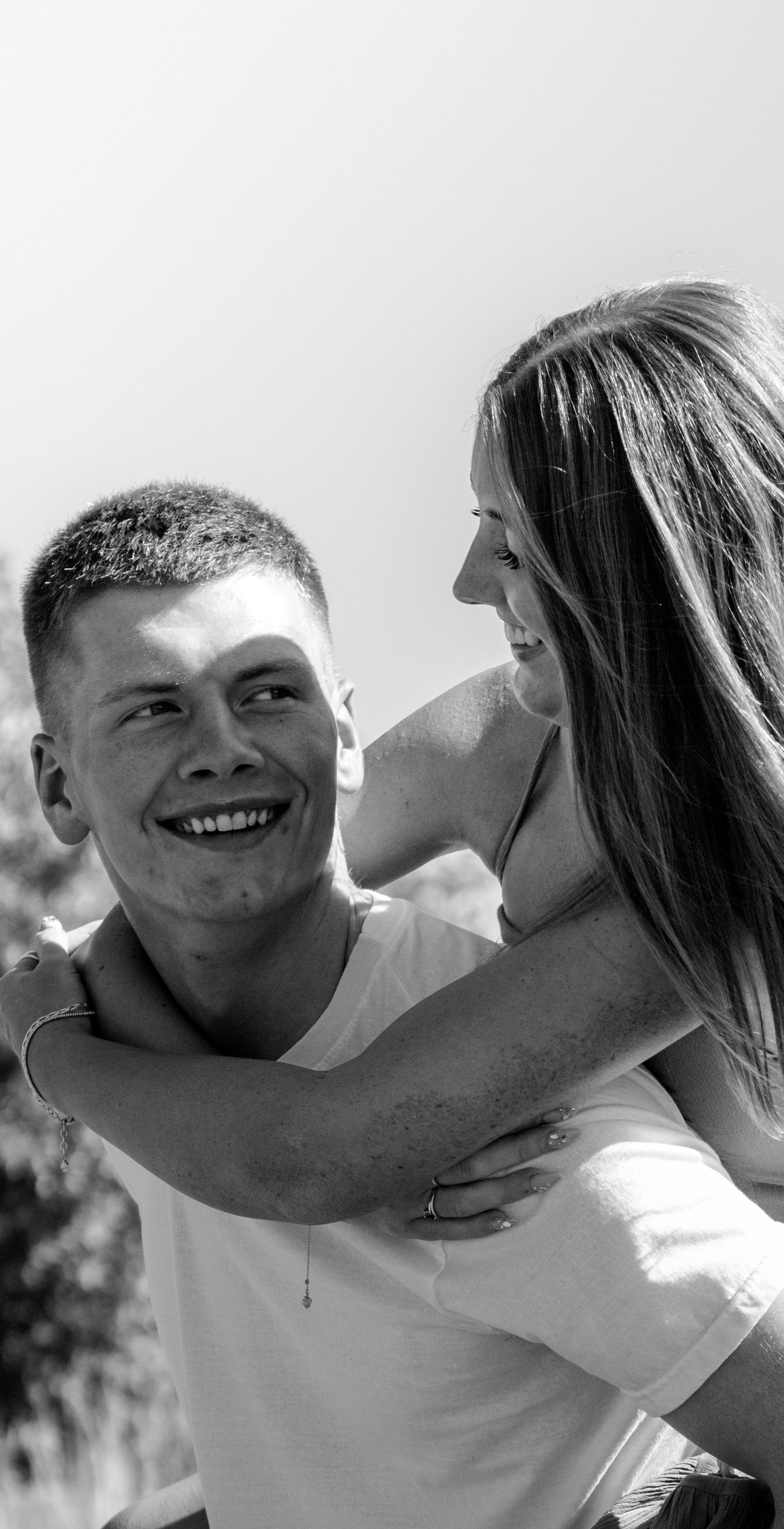 Black and white photo of a smiling young man giving a laughing woman a piggyback ride outdoors.
