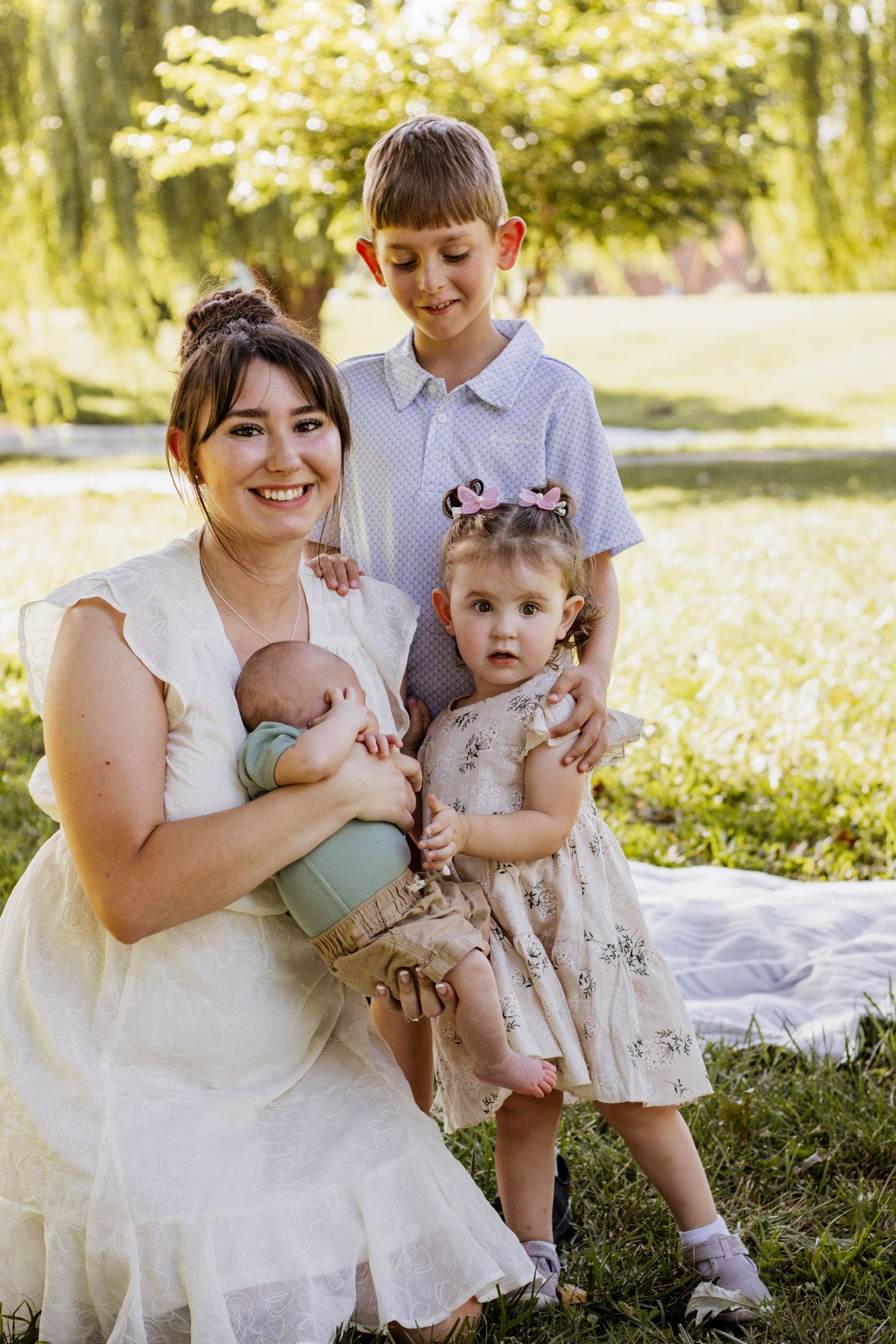 A smiling mother in a white dress holds a newborn outdoors, surrounded by her young daughter and older son in a park.