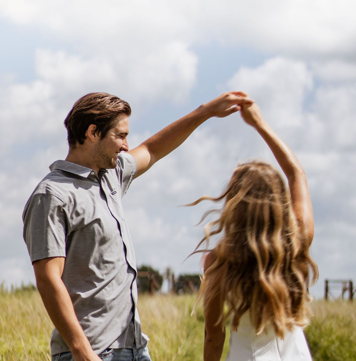 A young couple dances outdoors in a grassy field, man spinning woman under cloudy sky.
