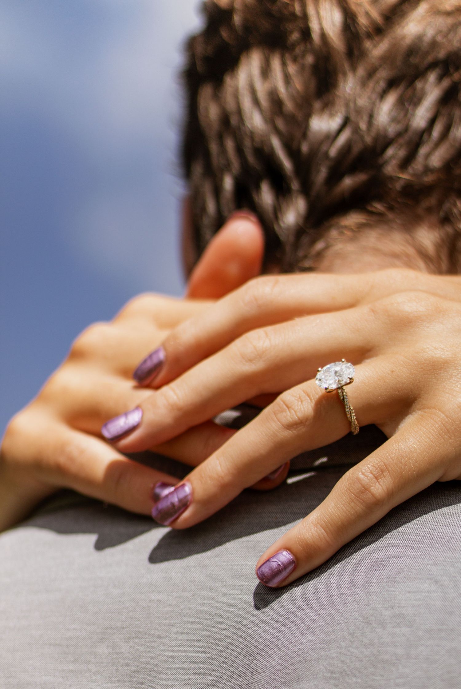 A woman with purple glitter nails showcases a stunning oval diamond engagement ring on a gold band outdoors.