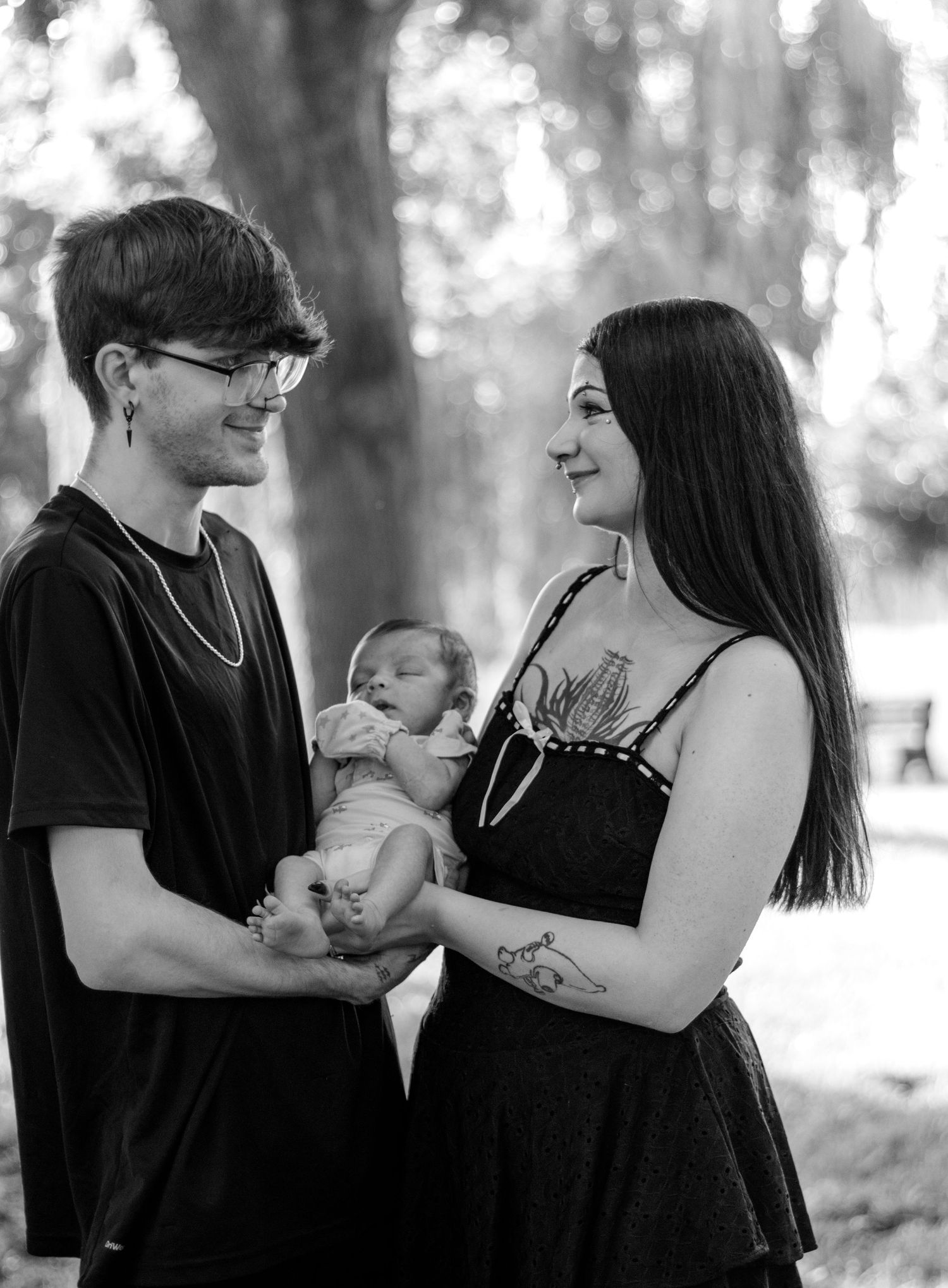 Black and white photo of a young couple smiling at each other while holding their newborn baby outdoors.