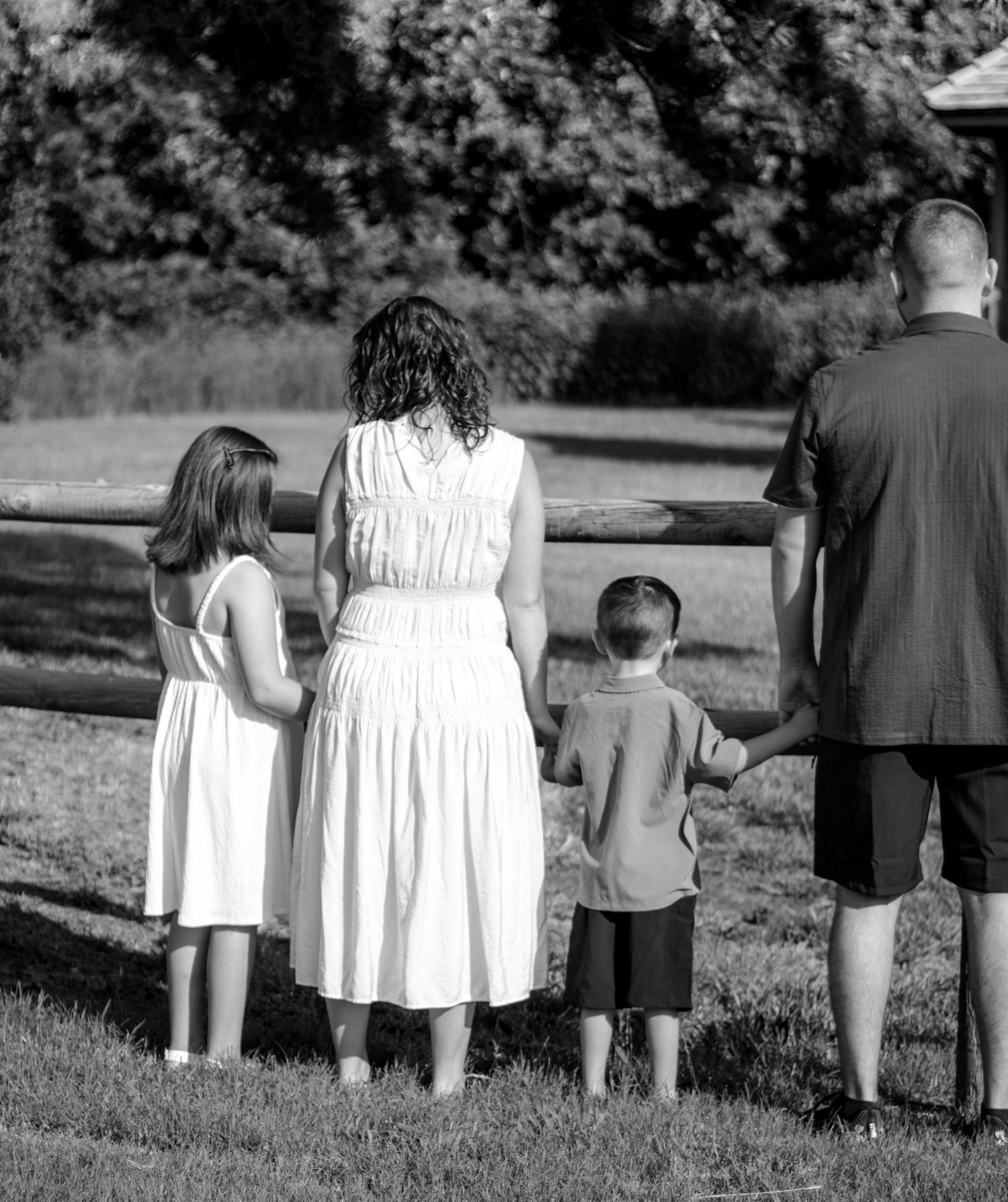 Black and white photo of a family of four holding hands while standing at a wooden fence outdoors.