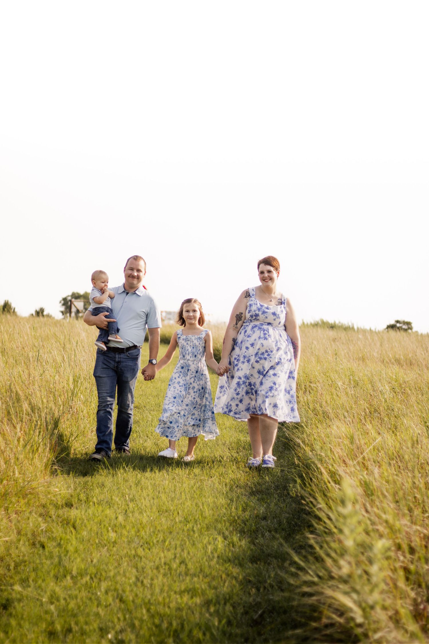 A happy family of four walks through a lush green field, with parents holding hands with their daughter while dad carries a toddler.
