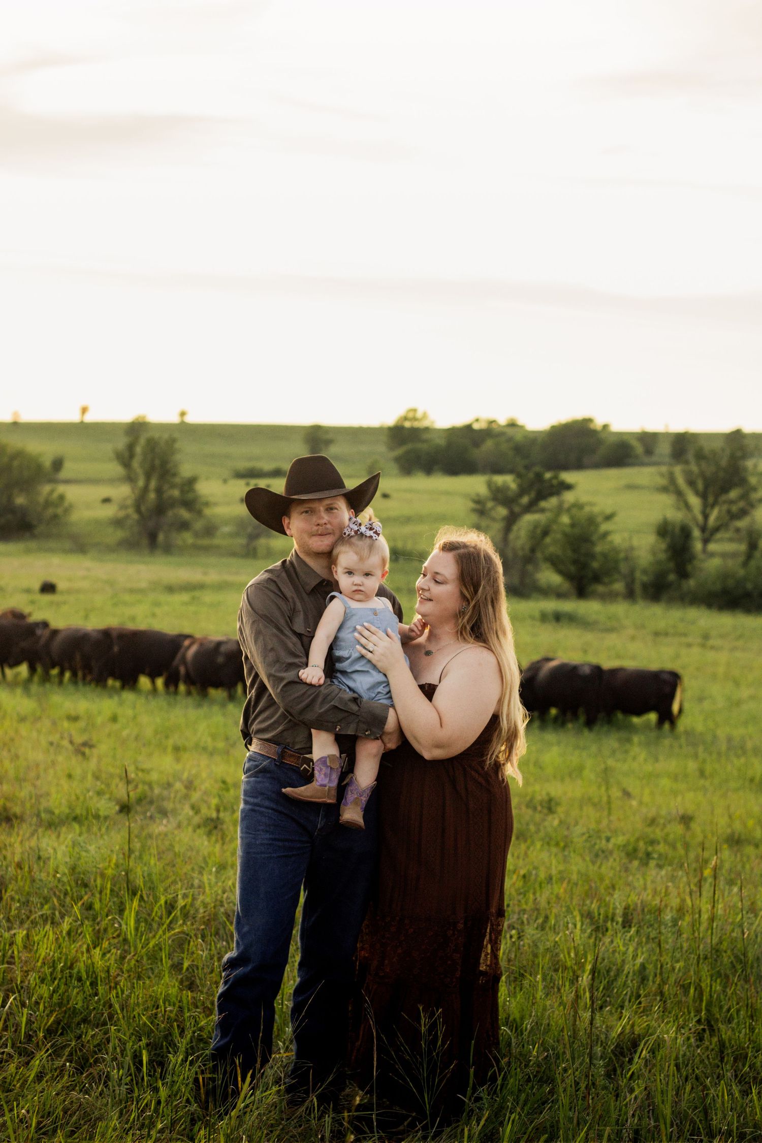 Young family with baby poses in green pasture at golden hour, cattle grazing in background, man in cowboy hat.