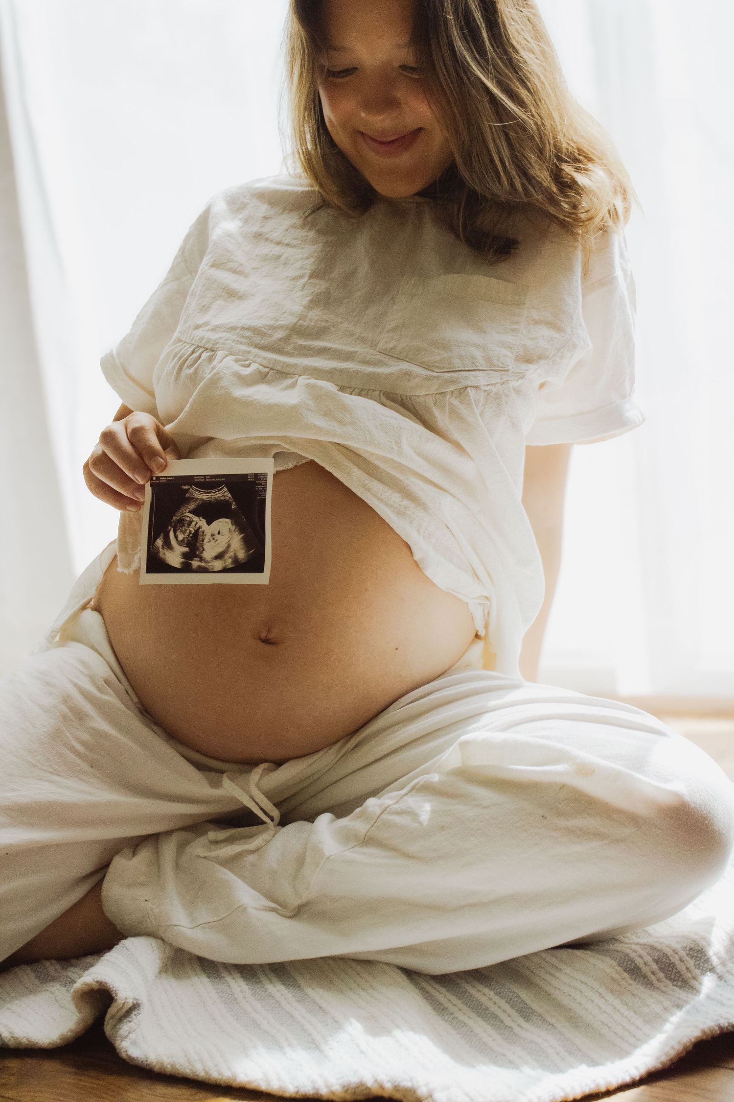 Smiling pregnant woman in white outfit holding ultrasound photo against her bare baby bump.