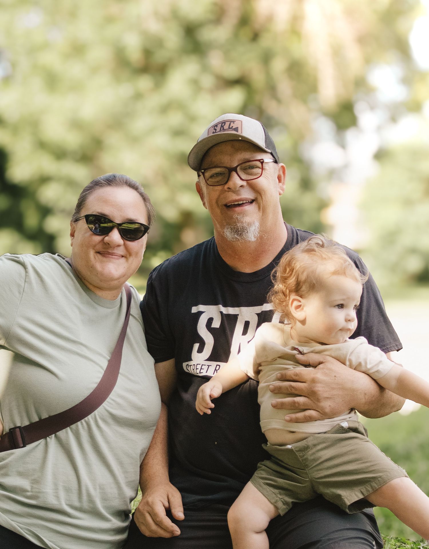 A smiling couple poses outdoors with a baby in a park setting, man wearing SRC Street Racing Channel shirt.