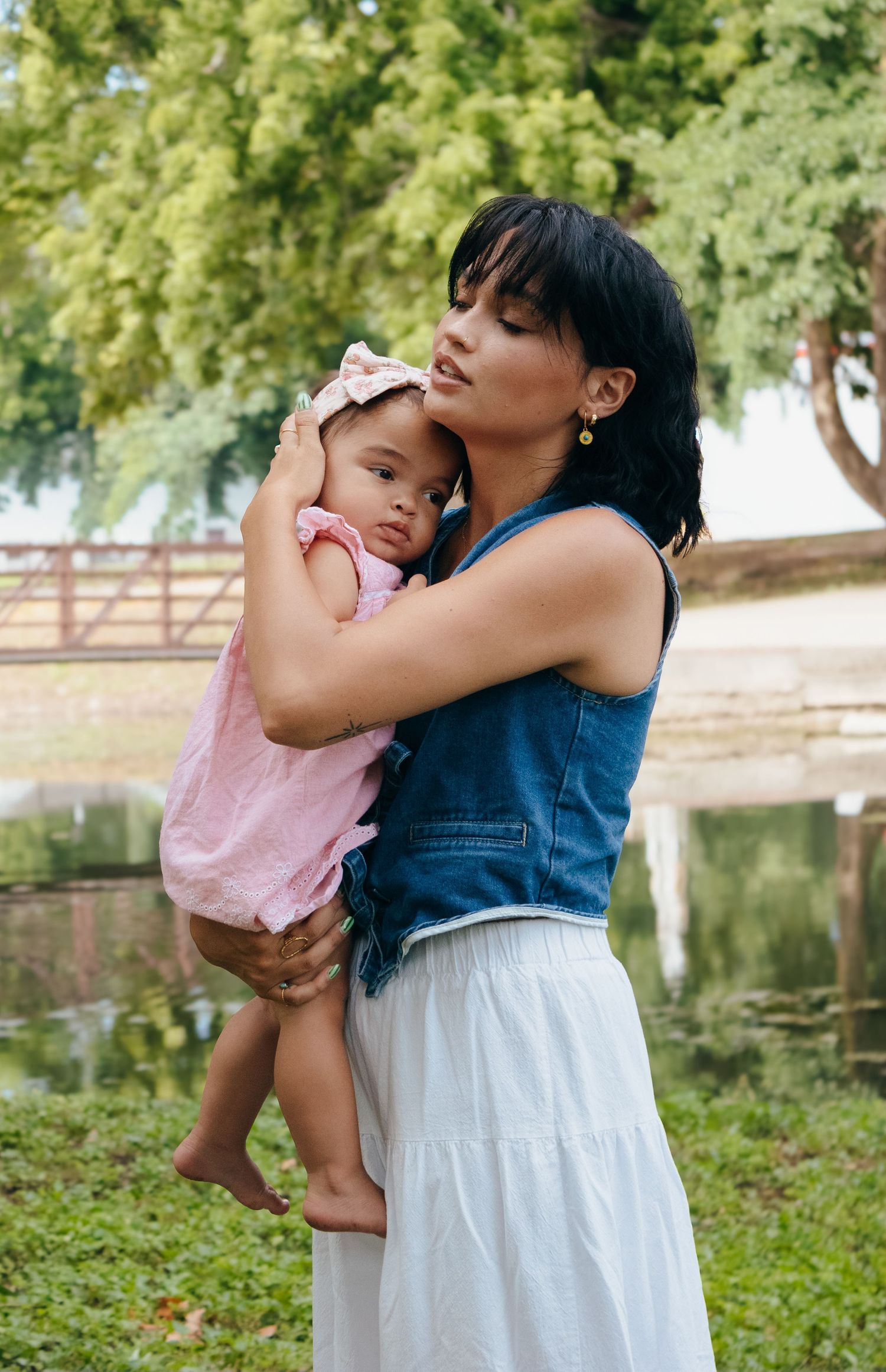 Loving mother in denim top holding young daughter in pink dress in park setting.