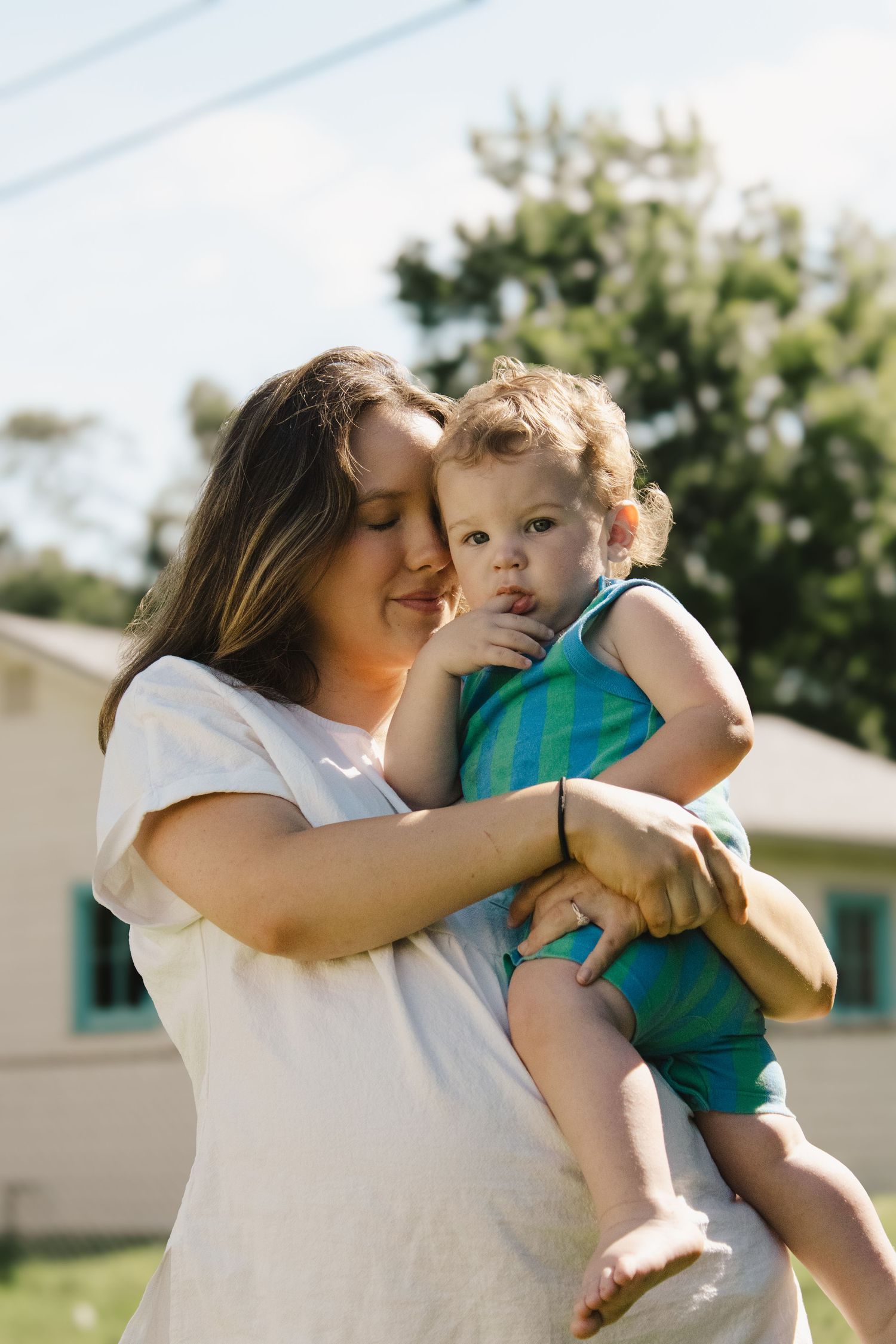 Young mother in white t-shirt lovingly holding toddler boy outdoors in sunny backyard.