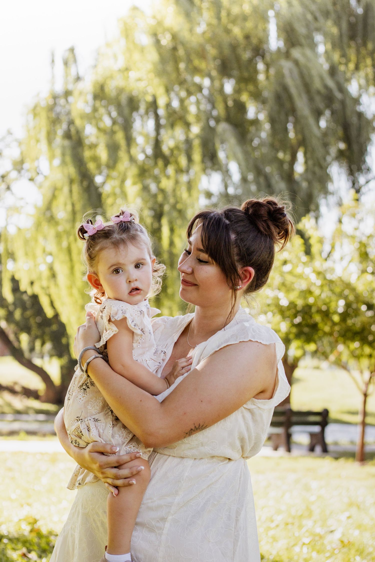 A young mother in a white dress holds her toddler daughter in a sunny park with a willow tree backdrop.