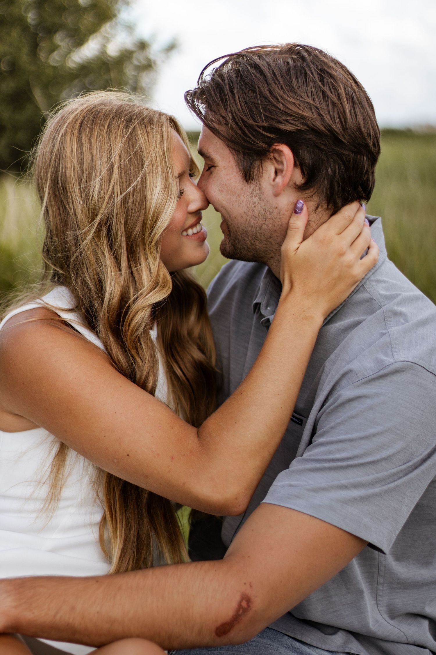 A couple shares a romantic moment outdoors, smiling and leaning in for a kiss, with a lush green background.