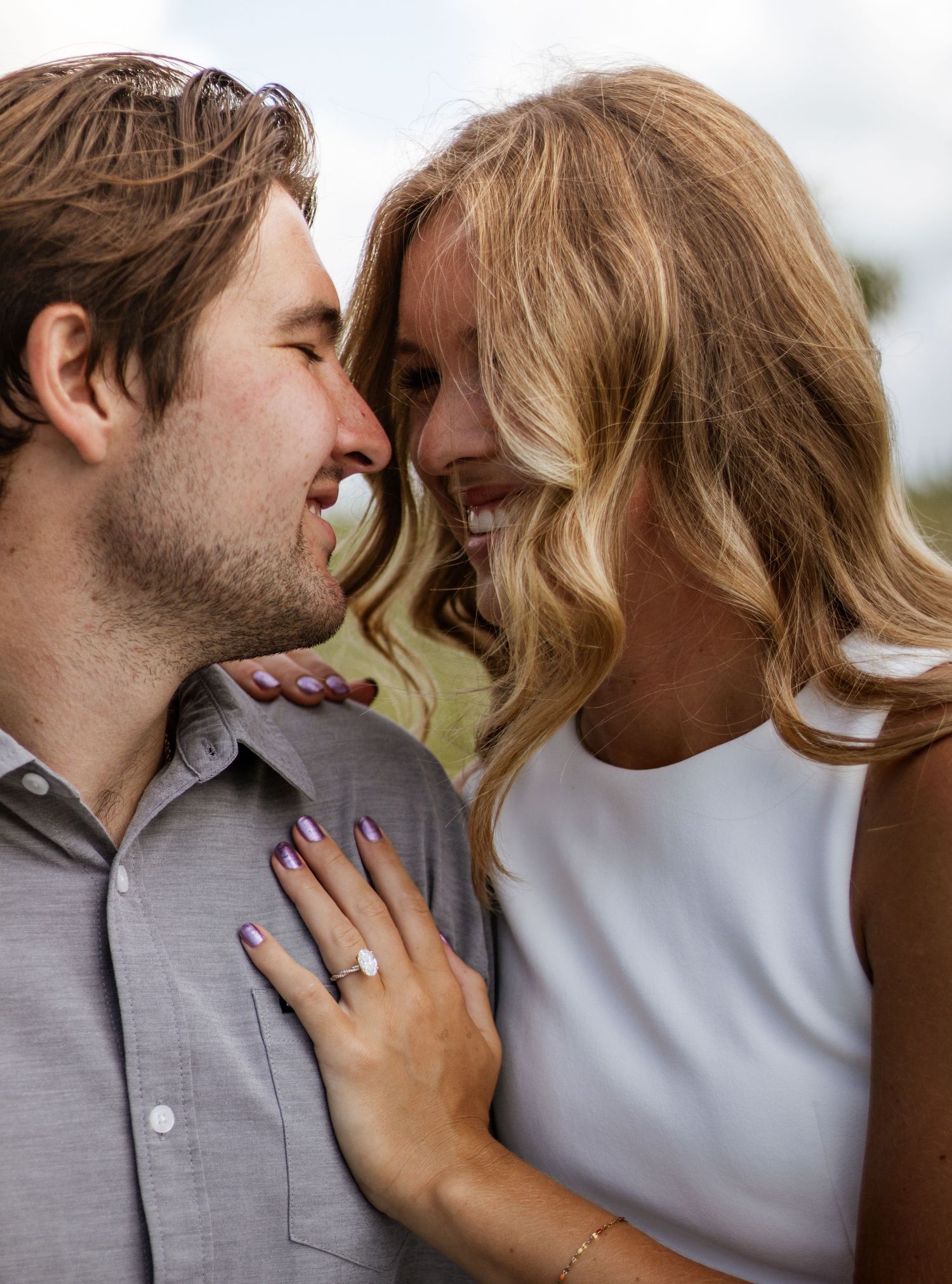 Smiling engaged couple nearly kissing, woman displaying diamond engagement ring on mans chest outdoors.