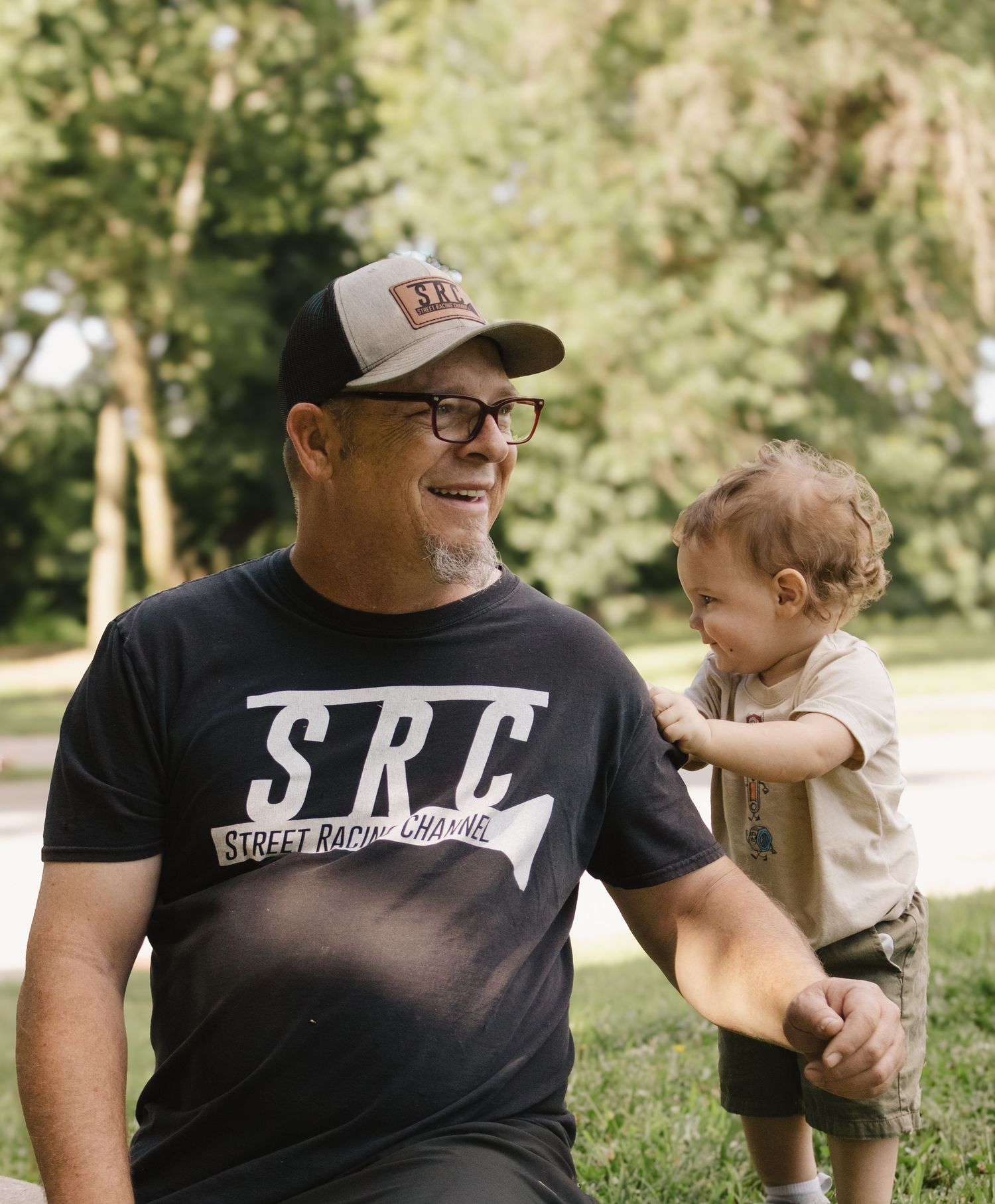 Man in SRC Street Racing Channel t-shirt smiling at a happy toddler in a sunny outdoor park.