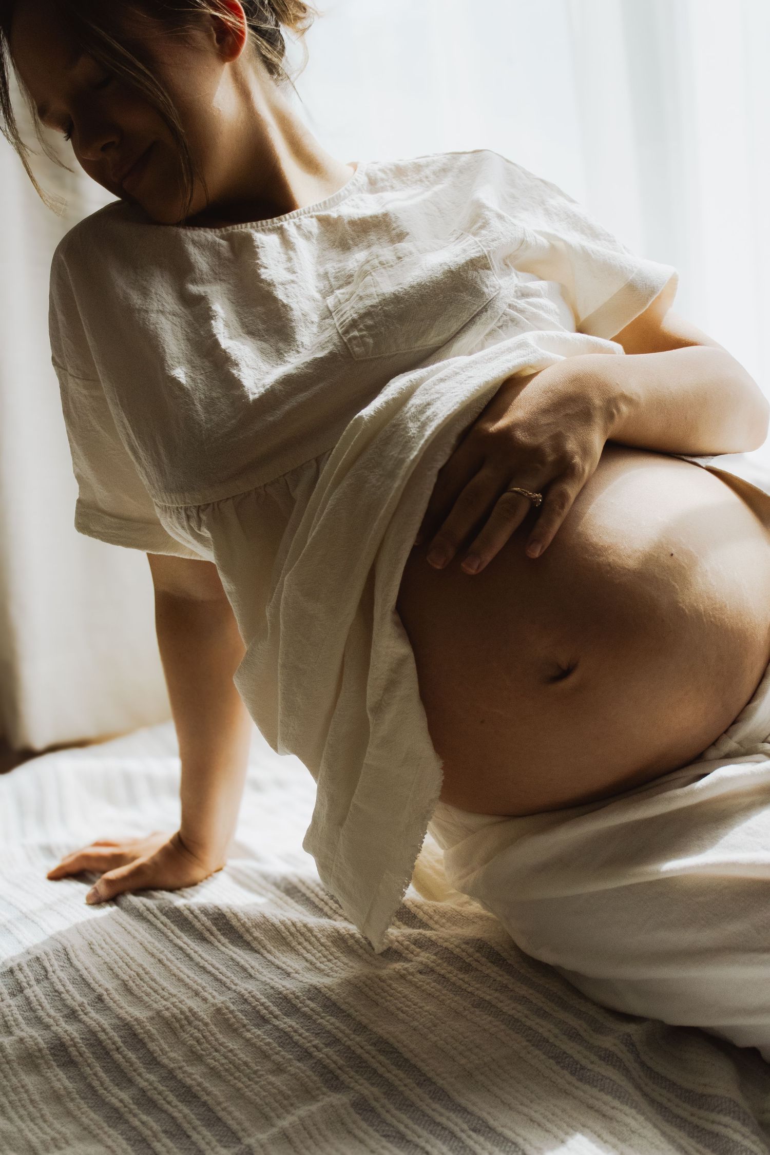 Pregnant woman in white linen top sitting on bed lifting shirt to reveal and hold her baby bump.