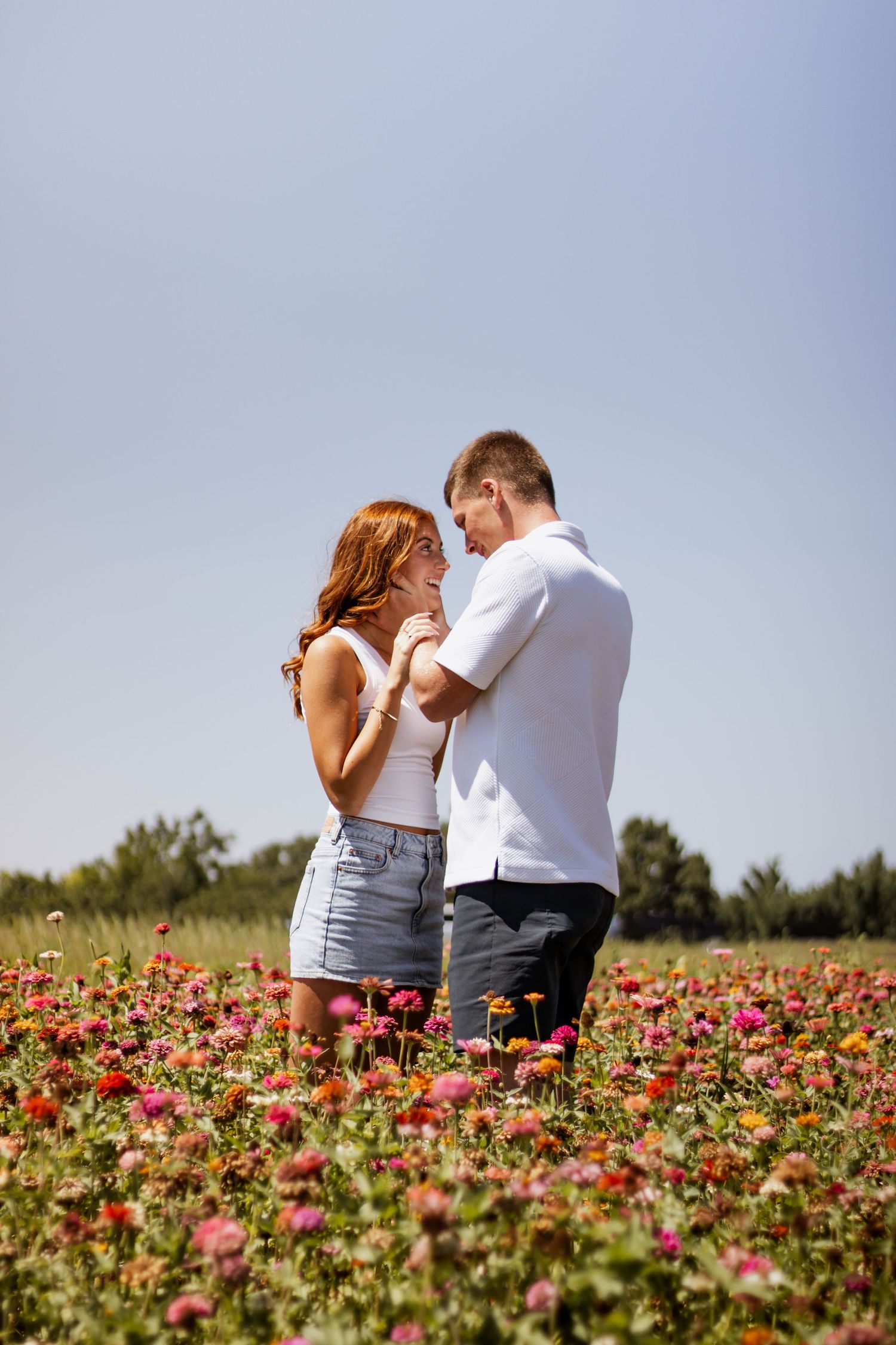 A romantic couple embracing in a vibrant flower field under a clear blue sky on a sunny day.