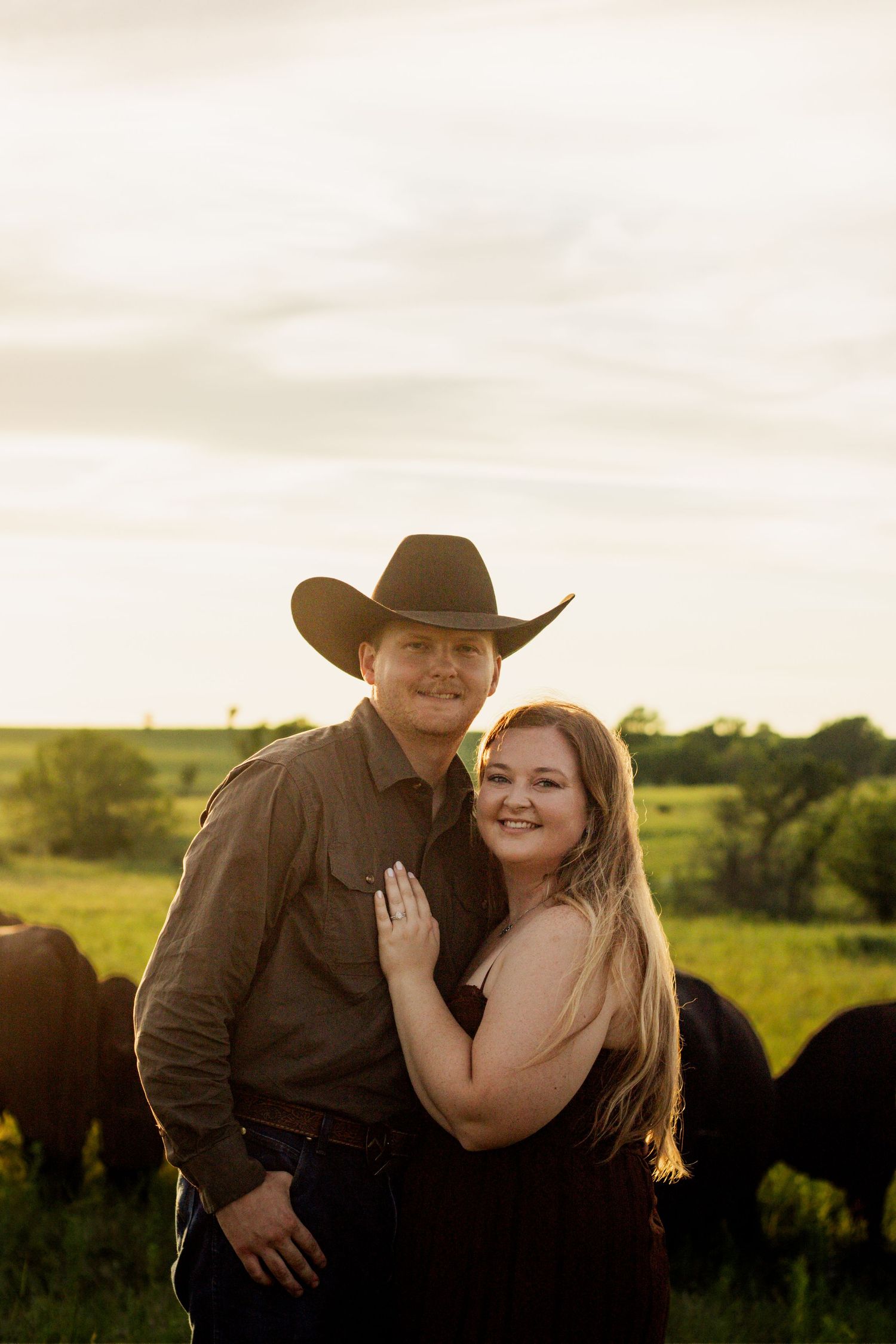 Couple embracing in a green pasture at golden hour, man wearing cowboy hat, cattle grazing in background.