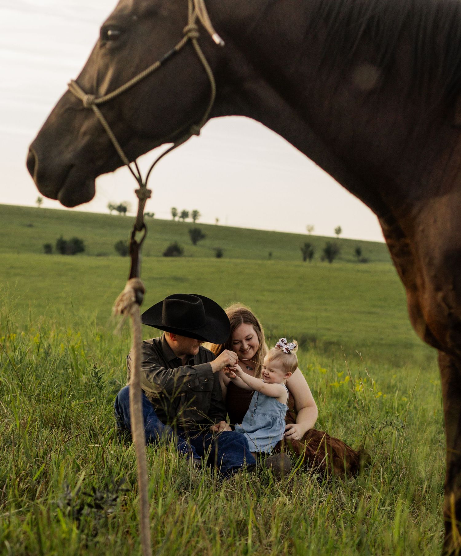 A family sits in a green field with a brown horse, a man in a cowboy hat, woman, and baby enjoying a countryside moment.