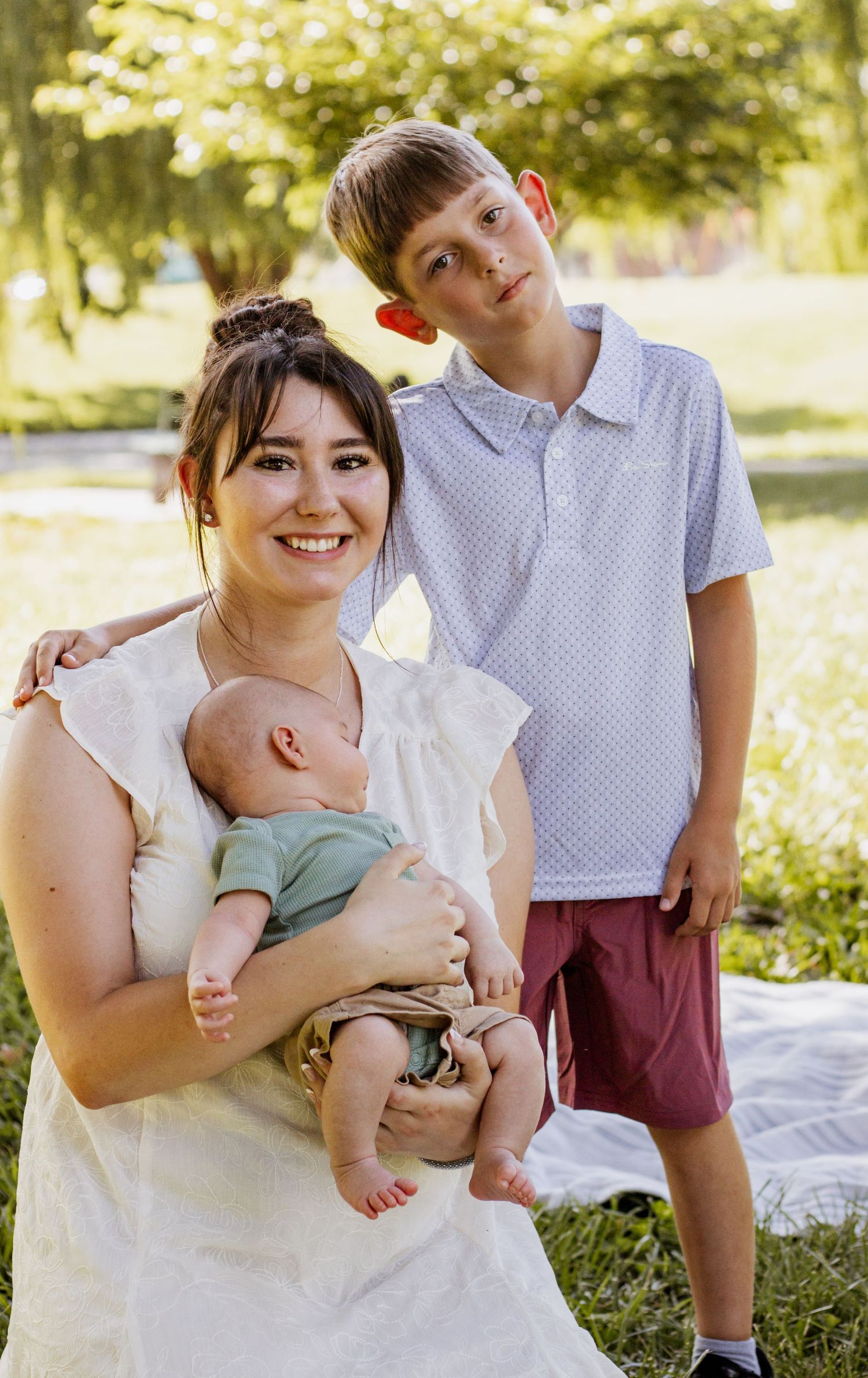 Smiling mother holding newborn baby outdoors with older boy standing beside her on green grass.