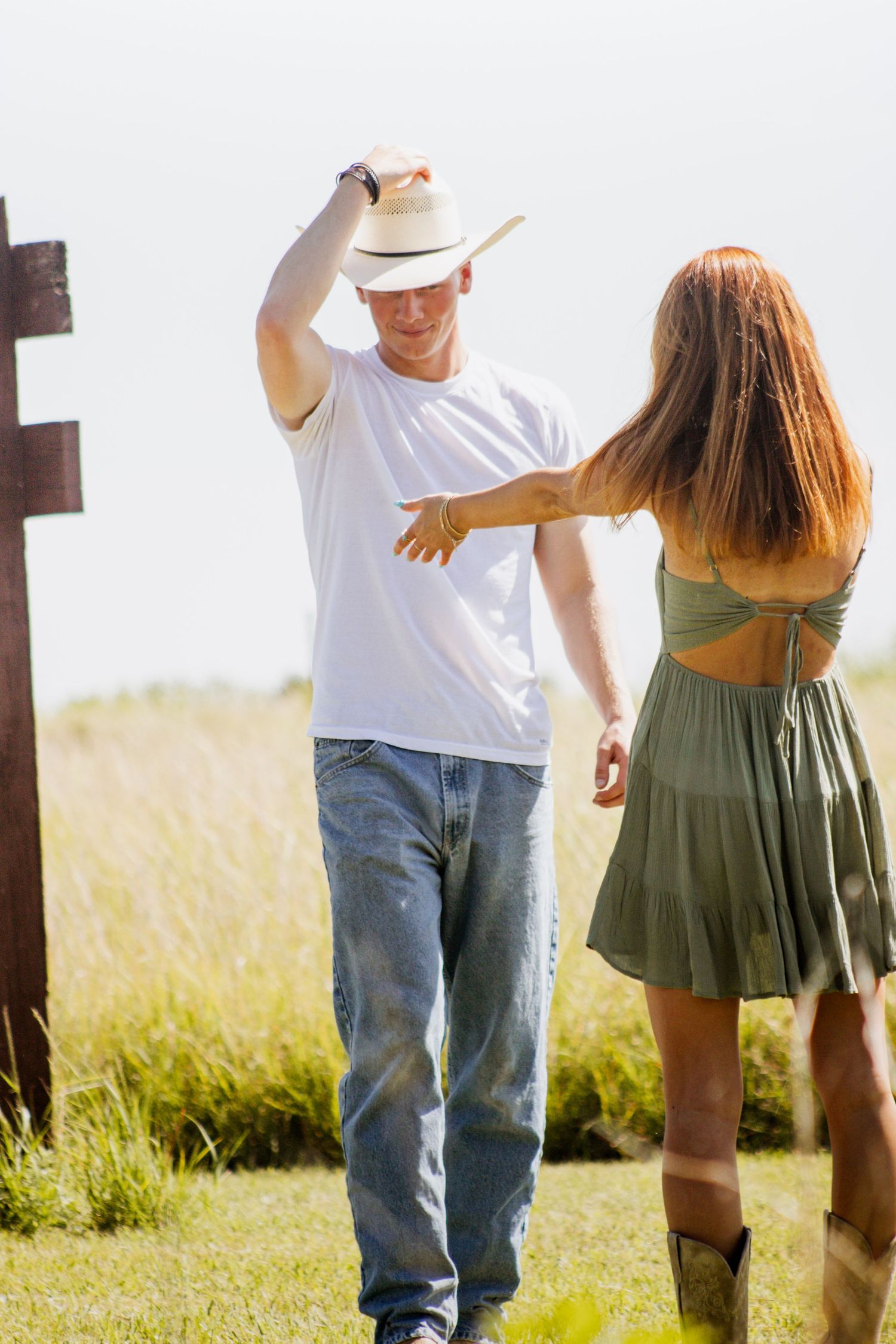 A man in a white t-shirt and cowboy hat dances with a woman in a green dress outdoors in a sunny field.