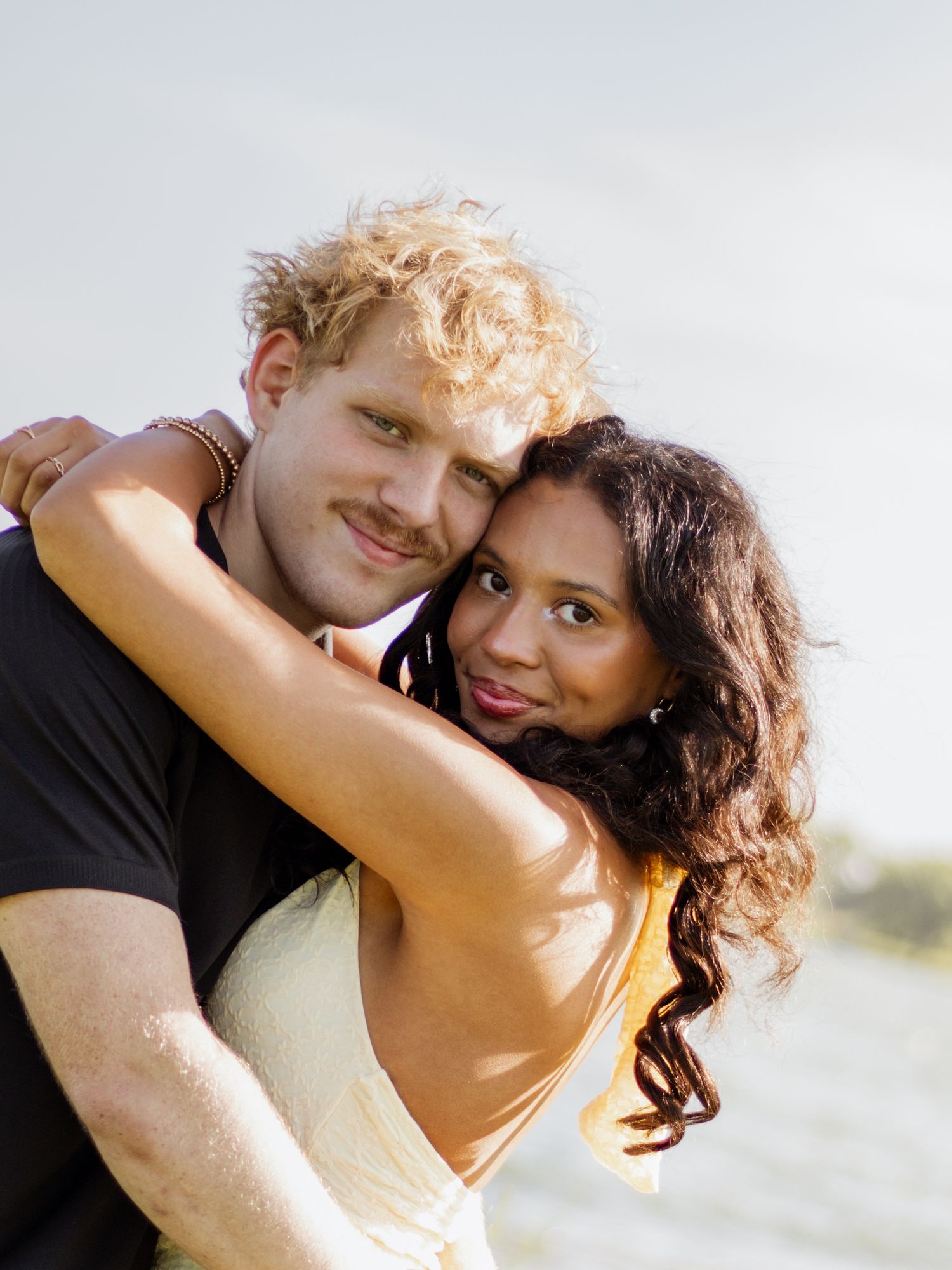 A smiling interracial couple embracing warmly outdoors near water, woman in yellow dress hugging man with curly blonde hair.