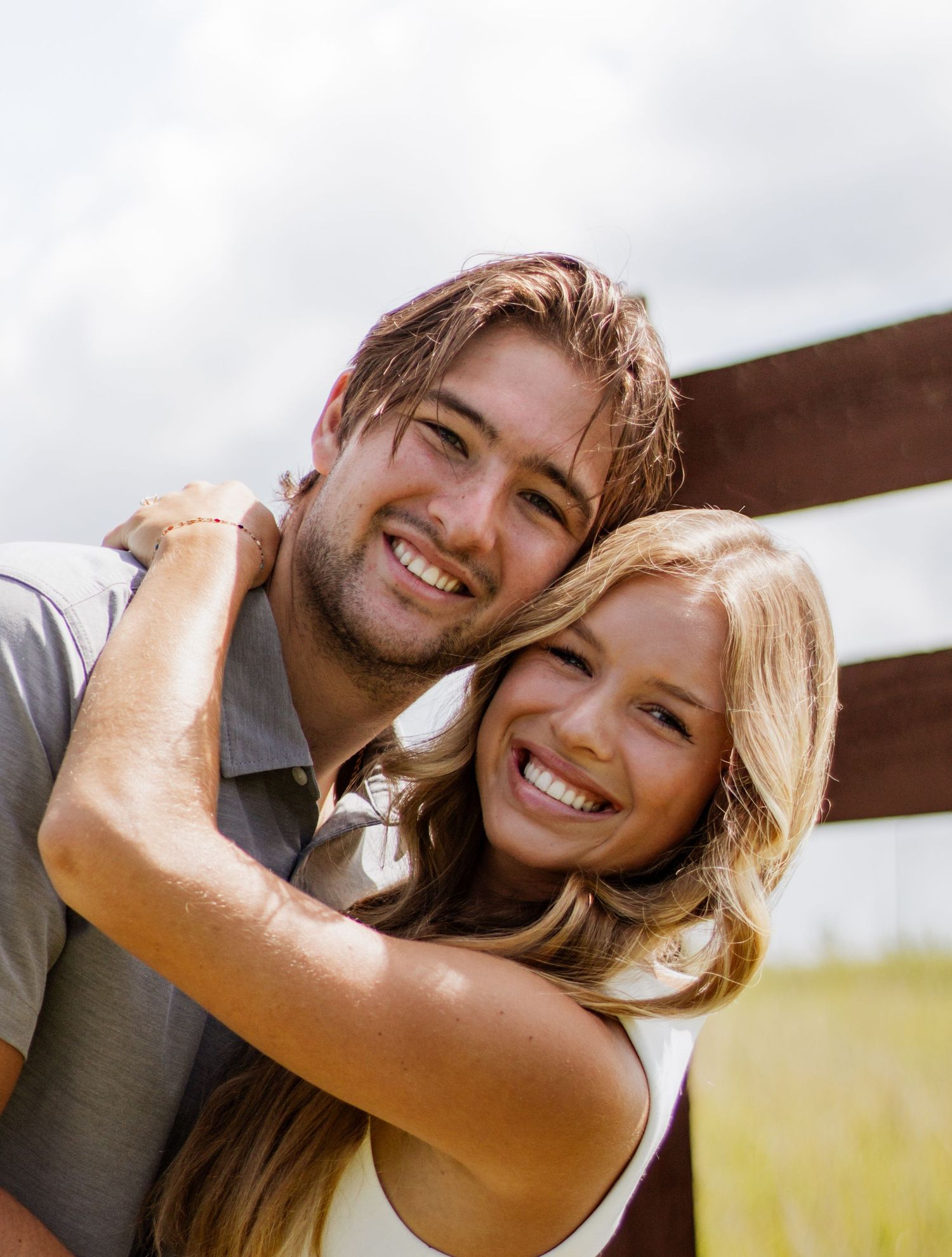 A smiling young couple embracing outdoors near a wooden fence, with the woman hugging the man from behind.