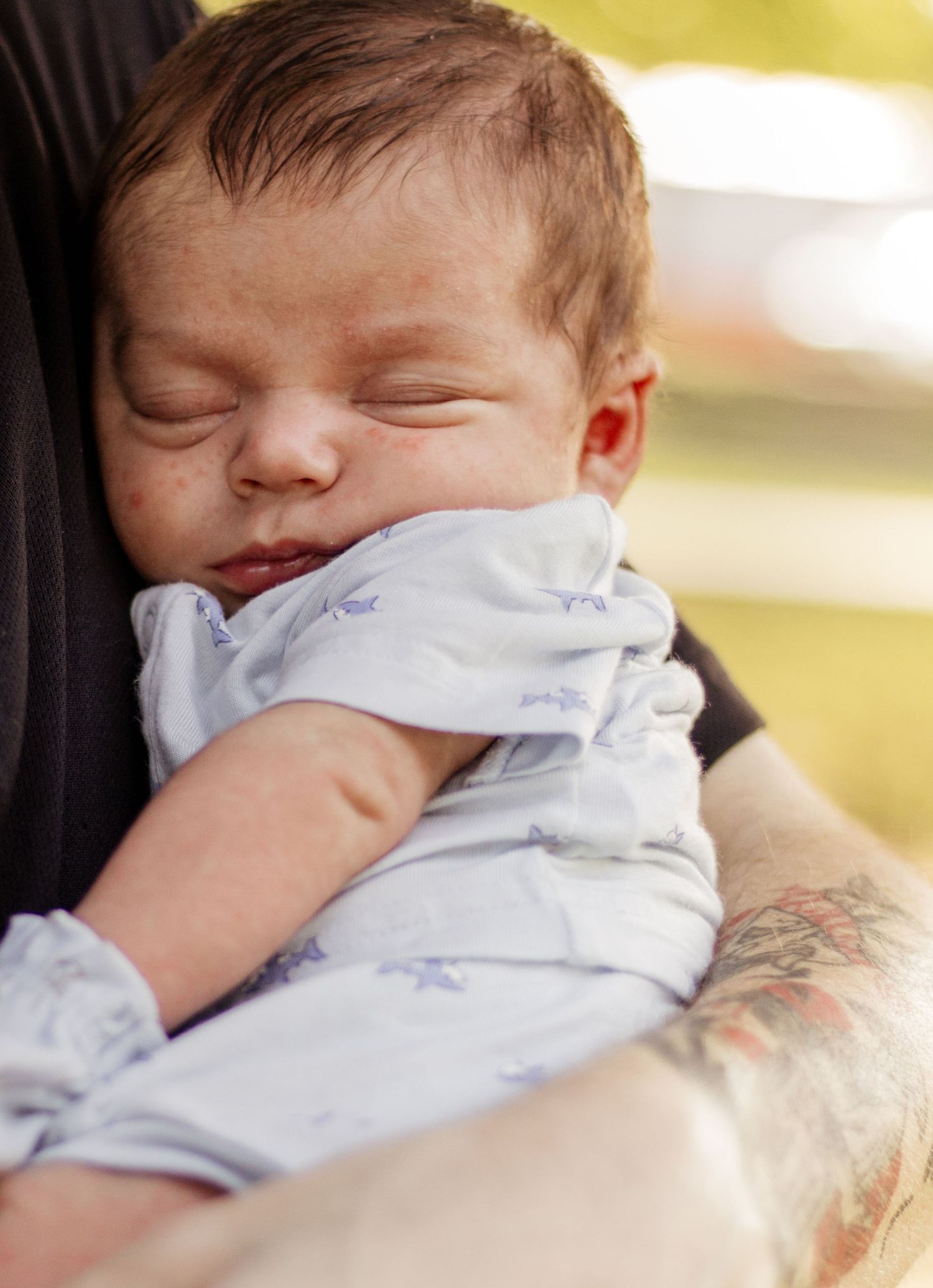 Newborn baby sleeping peacefully in the arms of a tattooed parent outdoors.