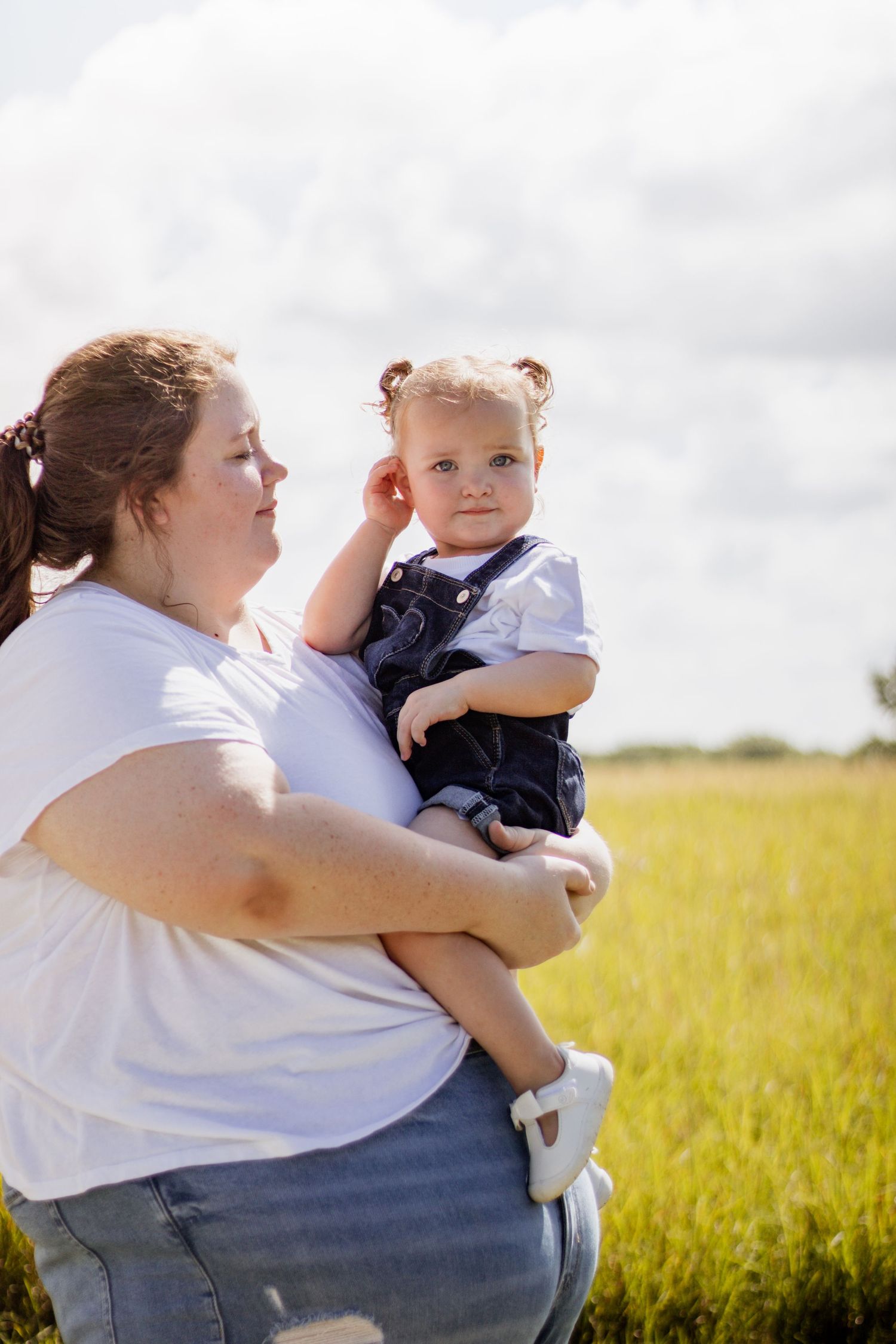 A mother holds her toddler in a sunny green field, both wearing casual clothes under a partly cloudy sky.