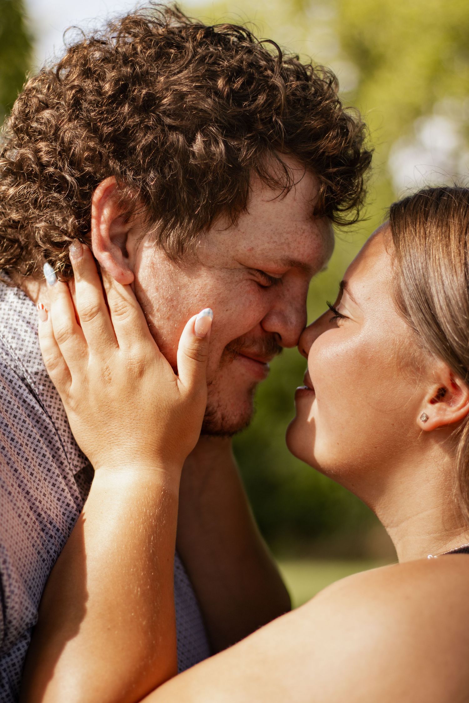 A romantic couple sharing an intimate moment outdoors, with the woman gently holding the man's face as they lean in to kiss.