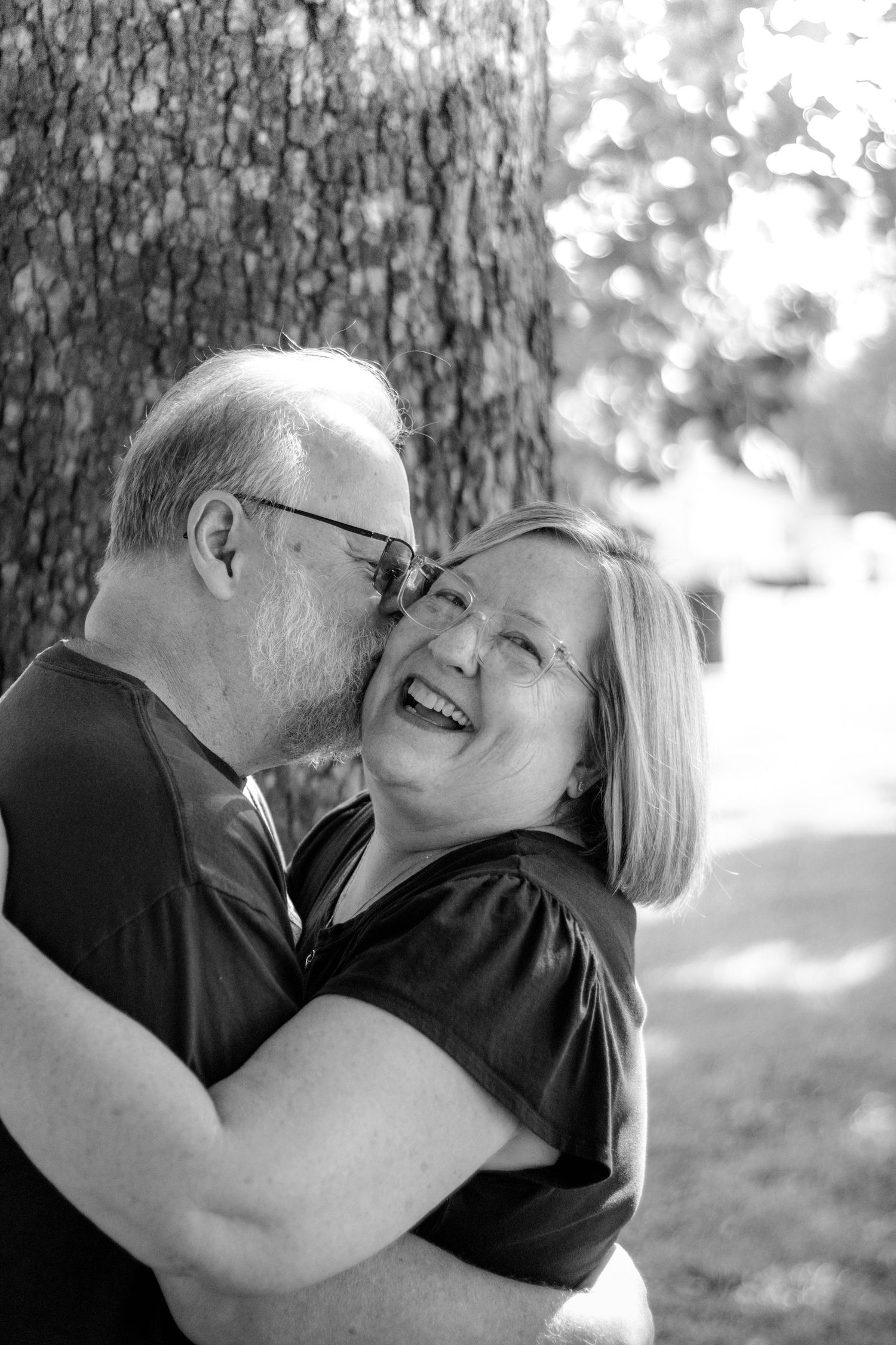 Black and white photo of an older couple hugging and laughing near a tree outdoors.