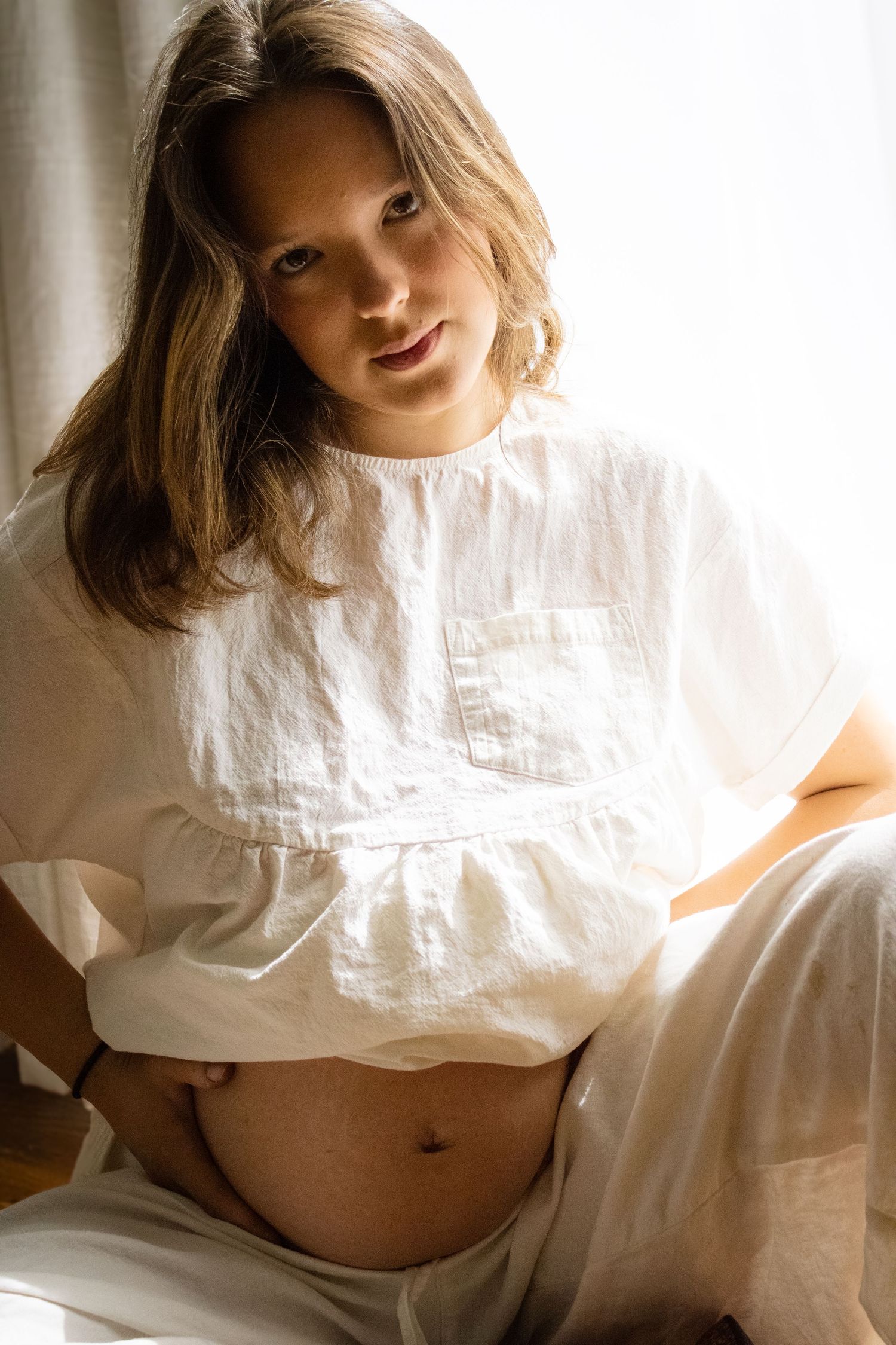 Pregnant woman in white linen top seated by window holding her bare belly.