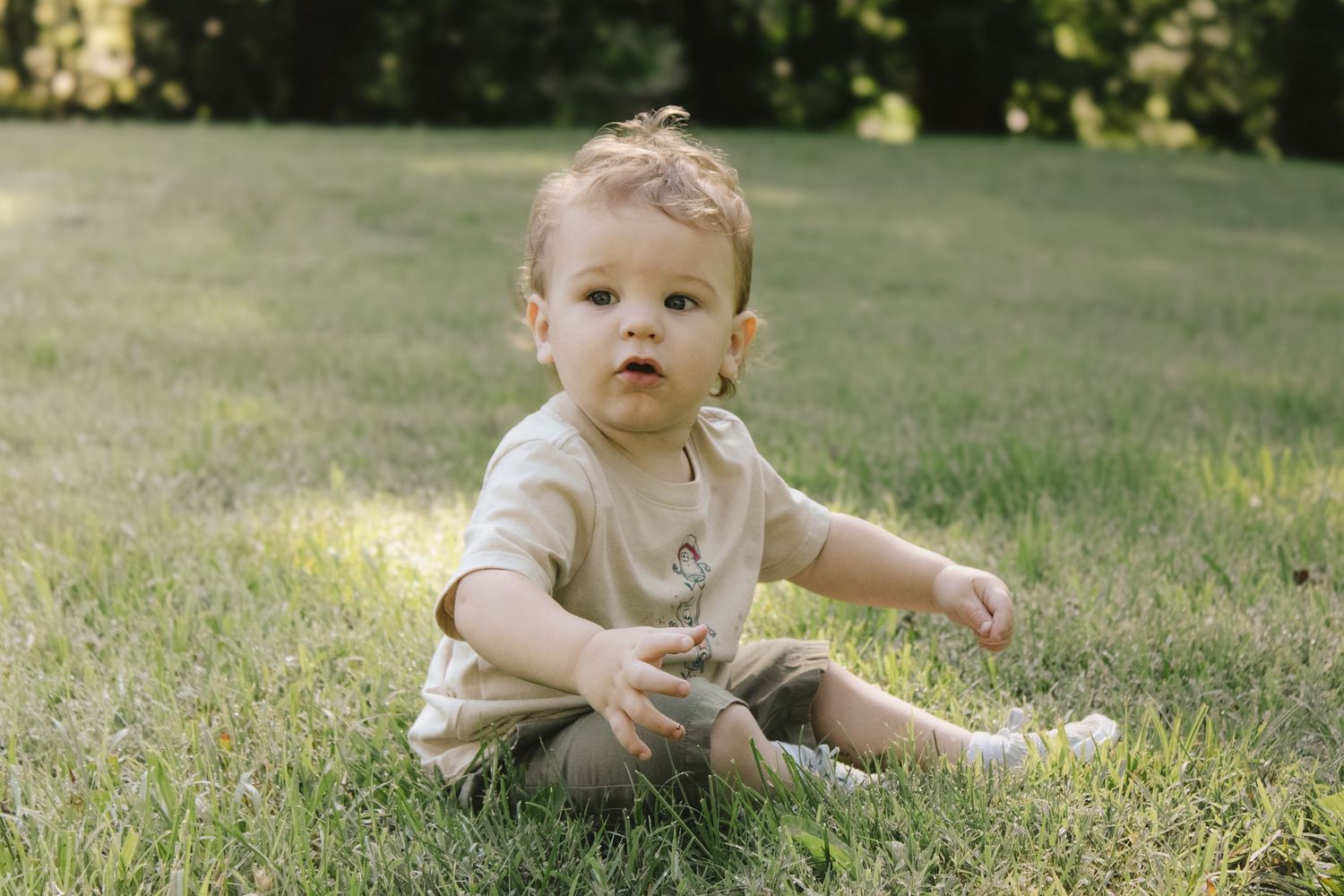 Adorable toddler in beige outfit sitting on green grass outdoors in warm sunlight.