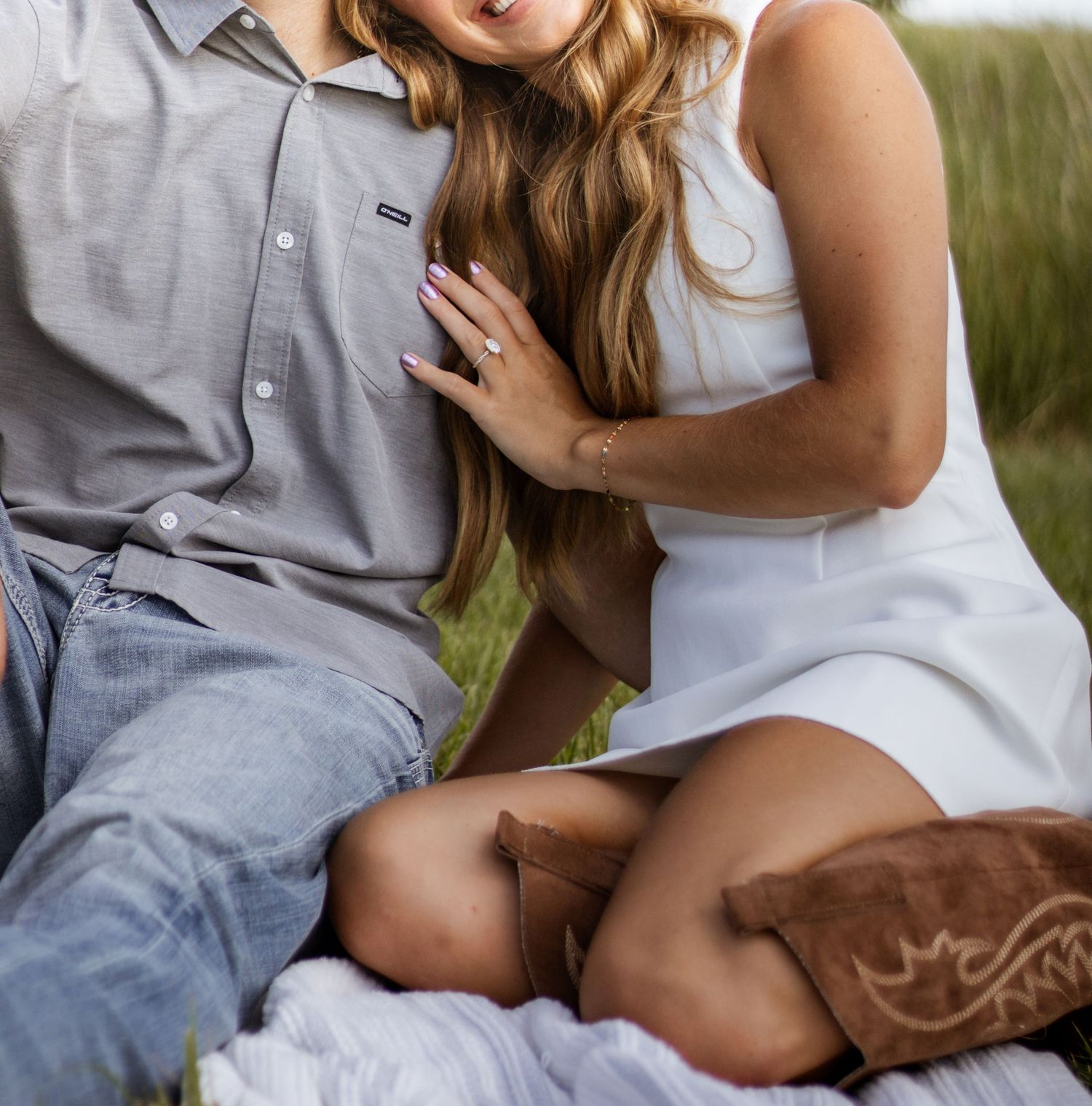 Couple sitting together outdoors, woman in white dress and cowboy boots, man in gray shirt and jeans.
