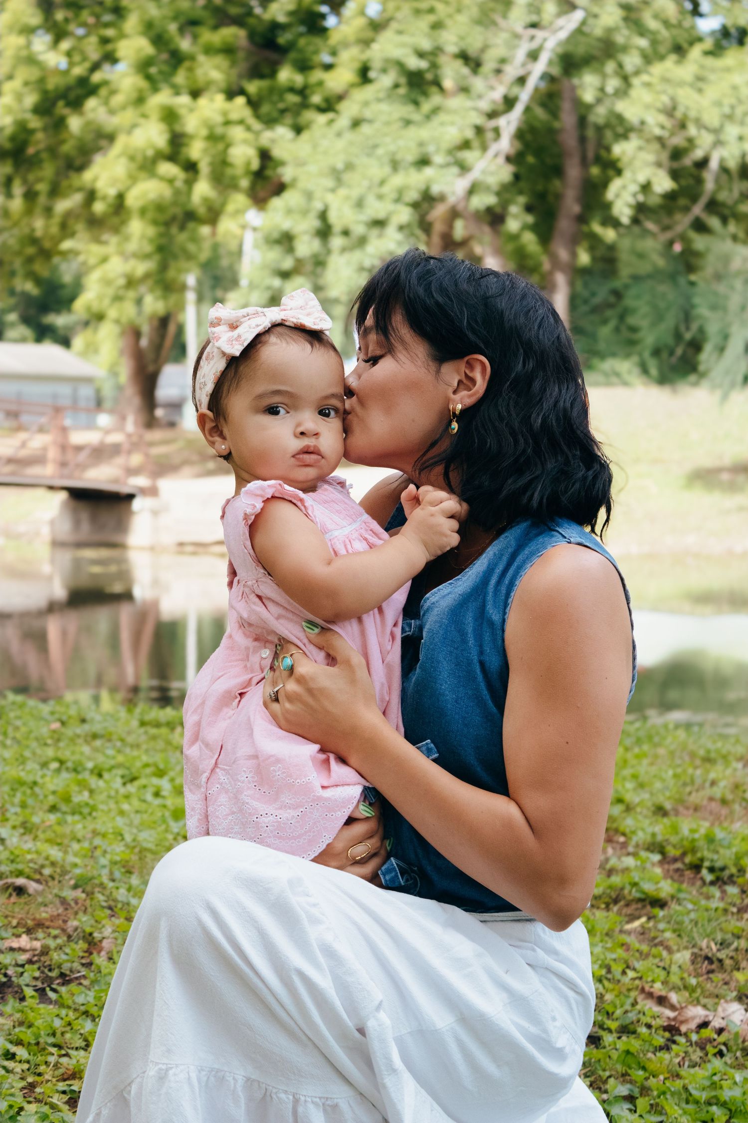 Mother kissing baby girl in pink dress and bow headband outdoors in green park setting.
