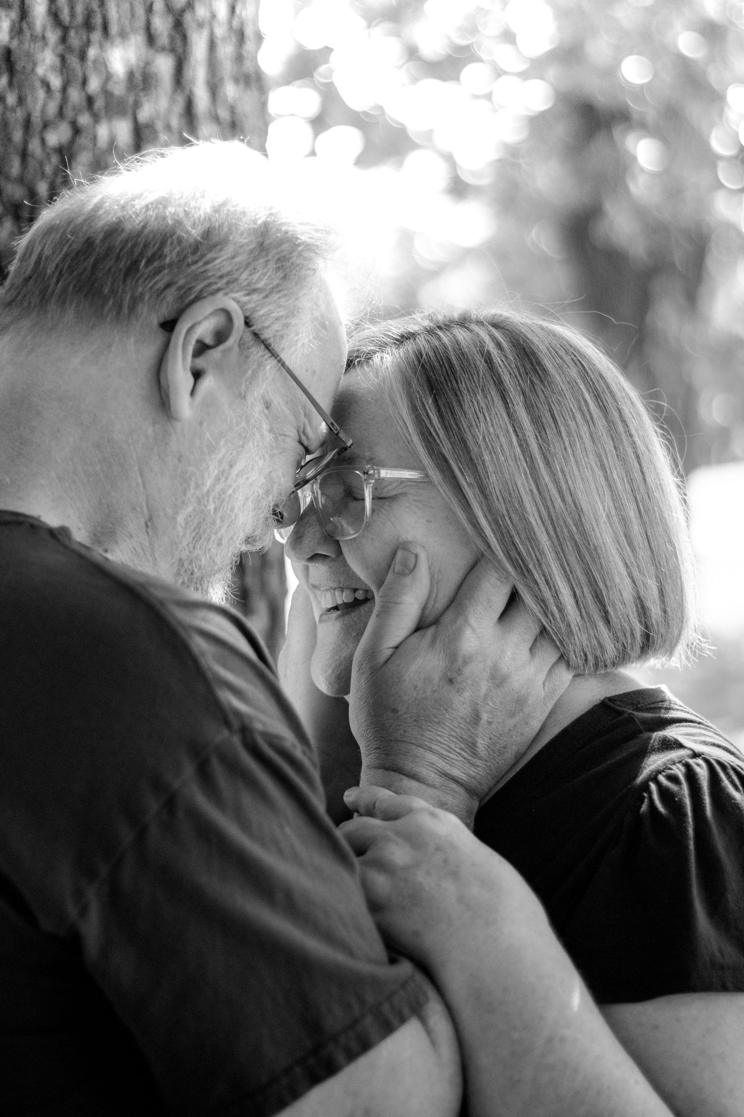 Black and white photo of an older couple sharing a tender moment, smiling and touching foreheads outdoors.
