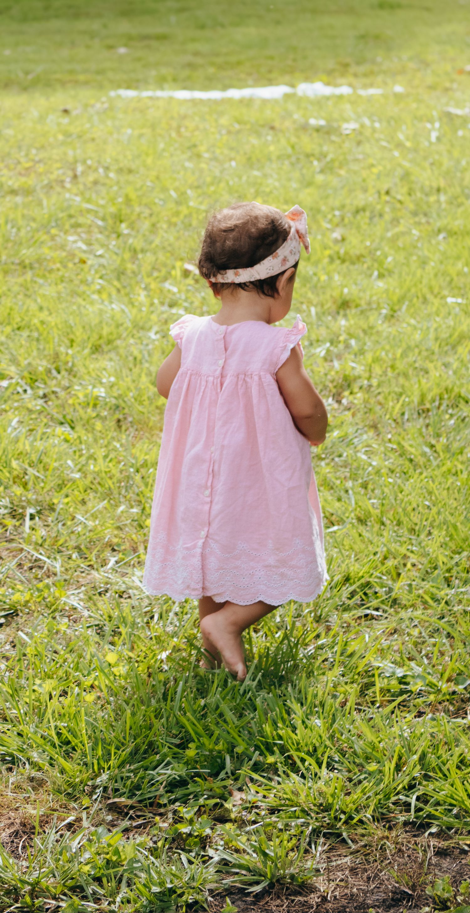 Toddler girl in pink dress and headband walking barefoot on lush green grass in park.