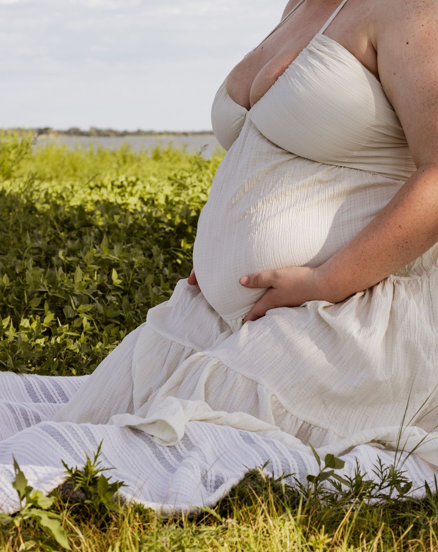 Pregnant woman in white dress sits on blanket in green field, cradling baby bump during maternity shoot.