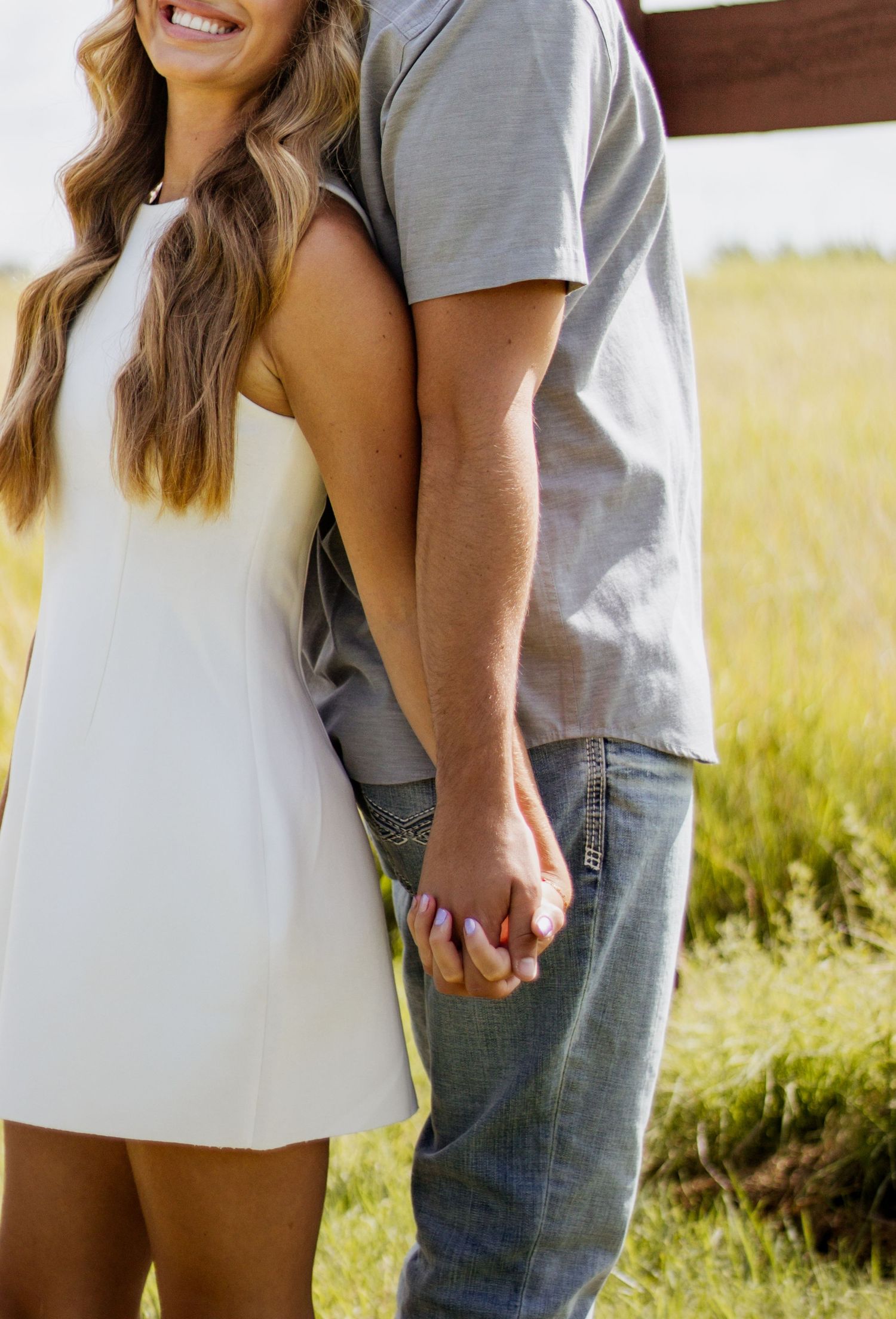 A couple holding hands in a green field, woman in white dress and man in gray shirt and jeans.