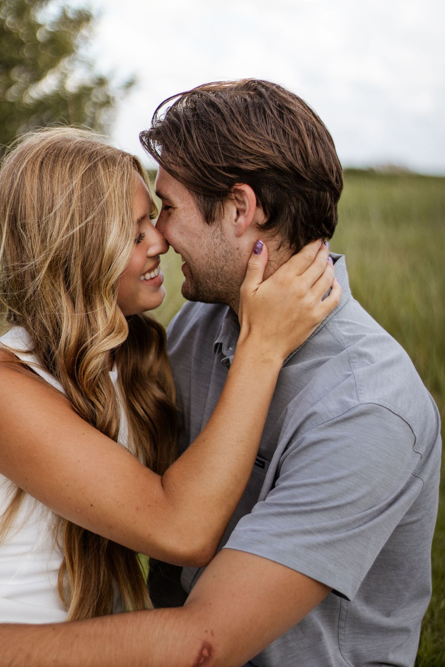 Romantic couple about to kiss outdoors, blonde woman holding mans face near green field.
