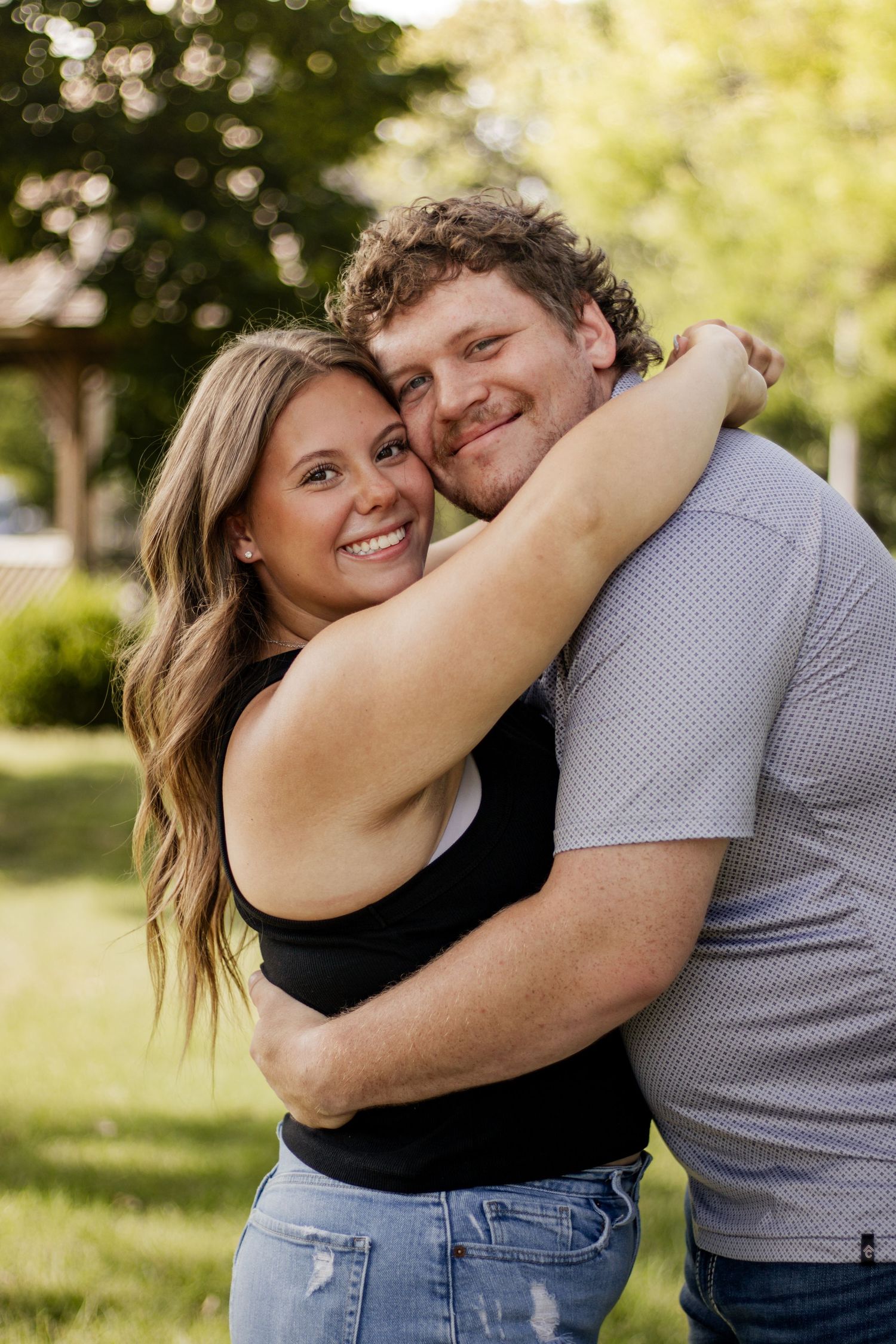 A smiling couple embraces outdoors in a sunny park, woman in black top and jeans hugging a curly-haired man in a gray shirt.