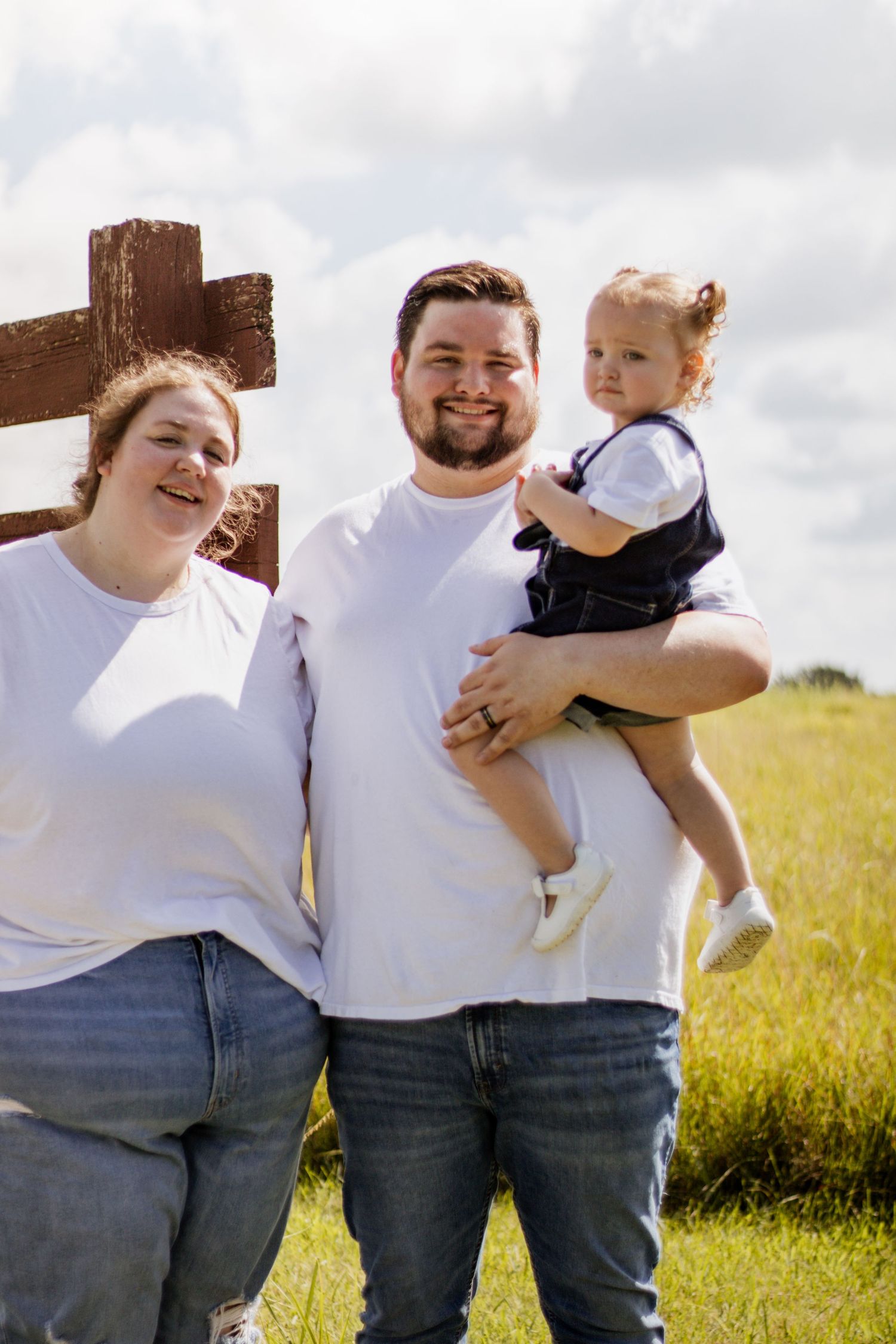 Family of three posing outdoors near wooden fence, parents in white tees and jeans, father holding toddler girl in denim overalls.