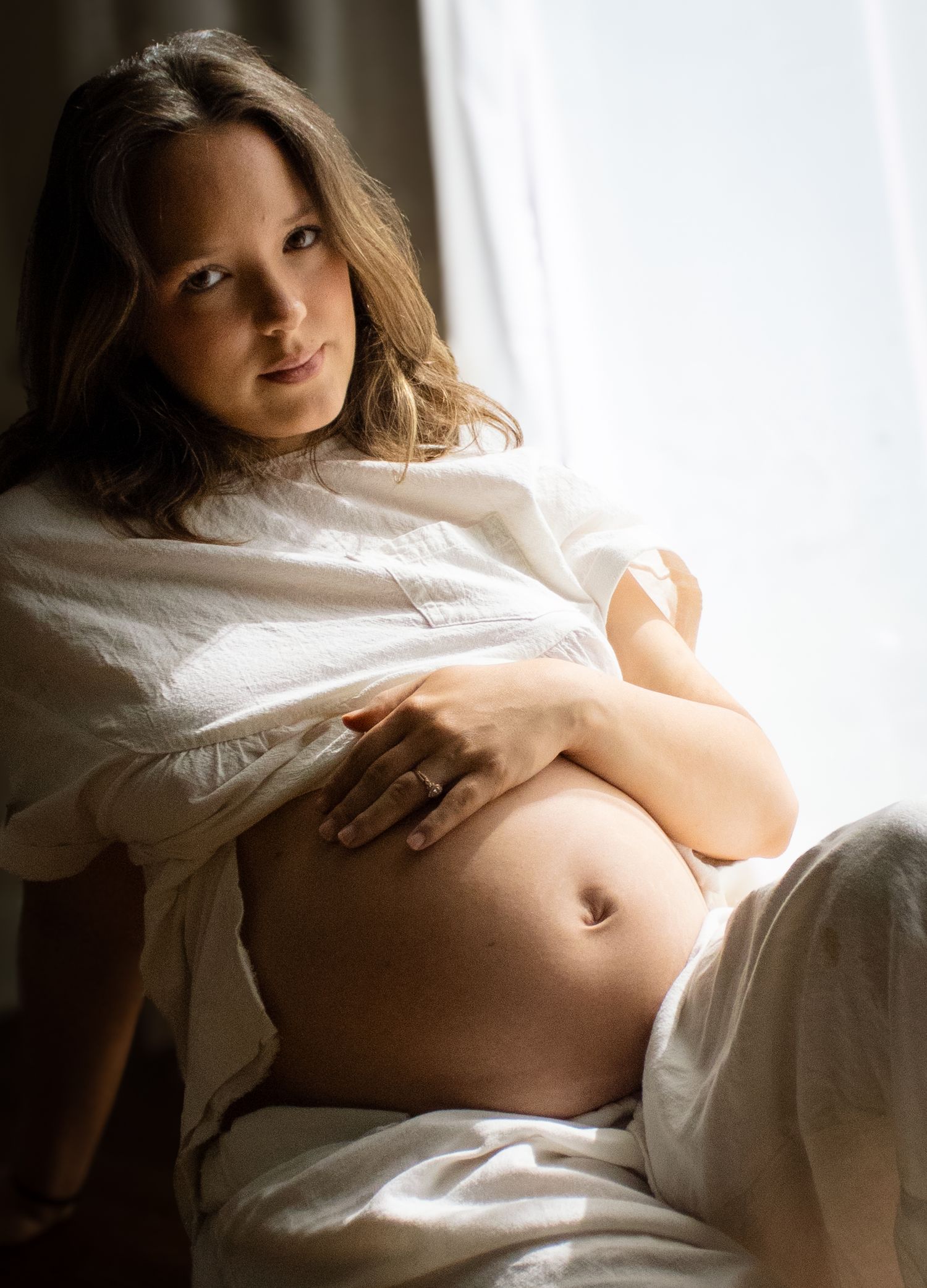 Pregnant woman in open white shirt sitting by window cradling her bare belly.