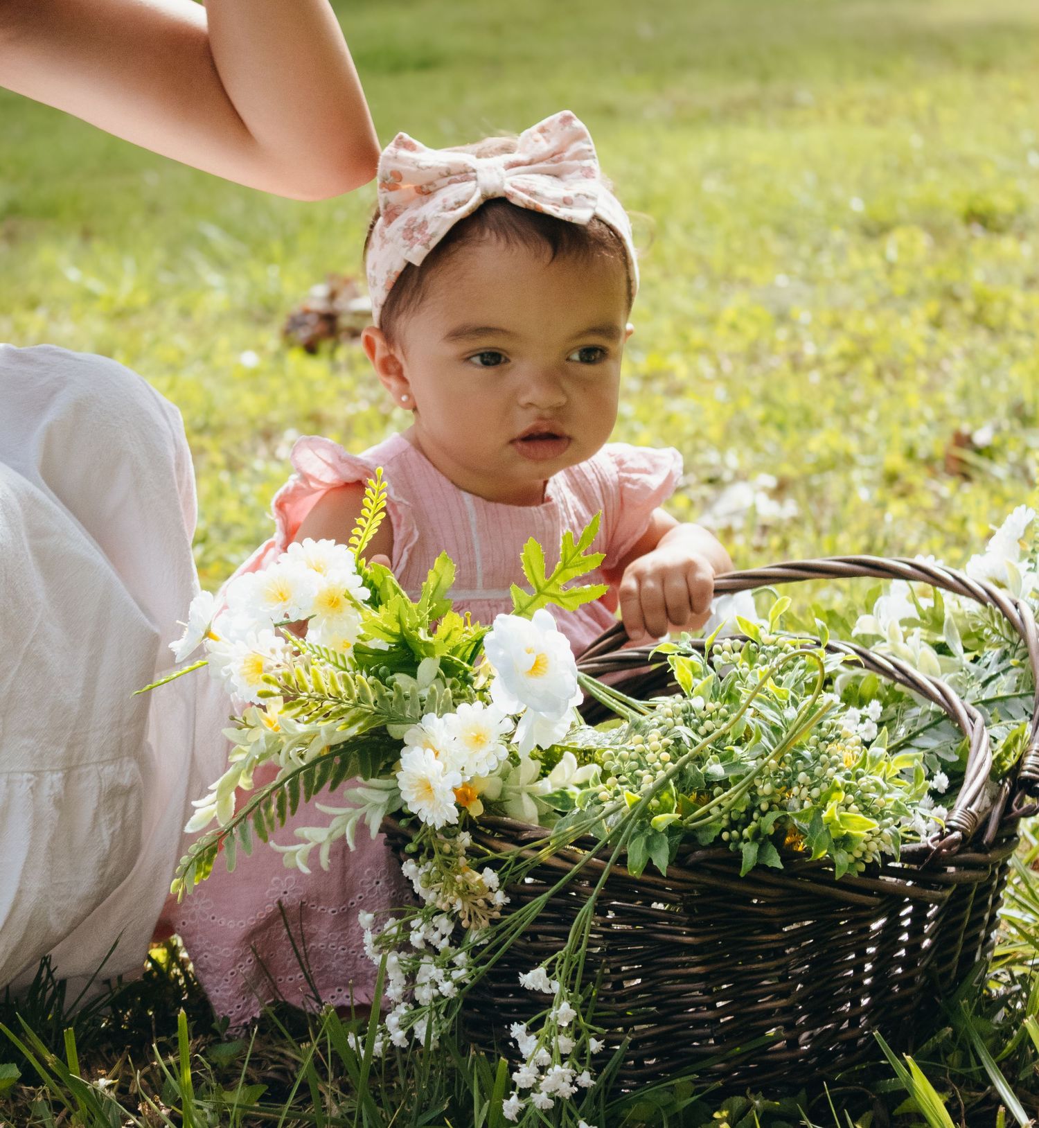 Adorable baby girl in pink dress and floral headband sitting in grass with a wicker basket of white flowers.