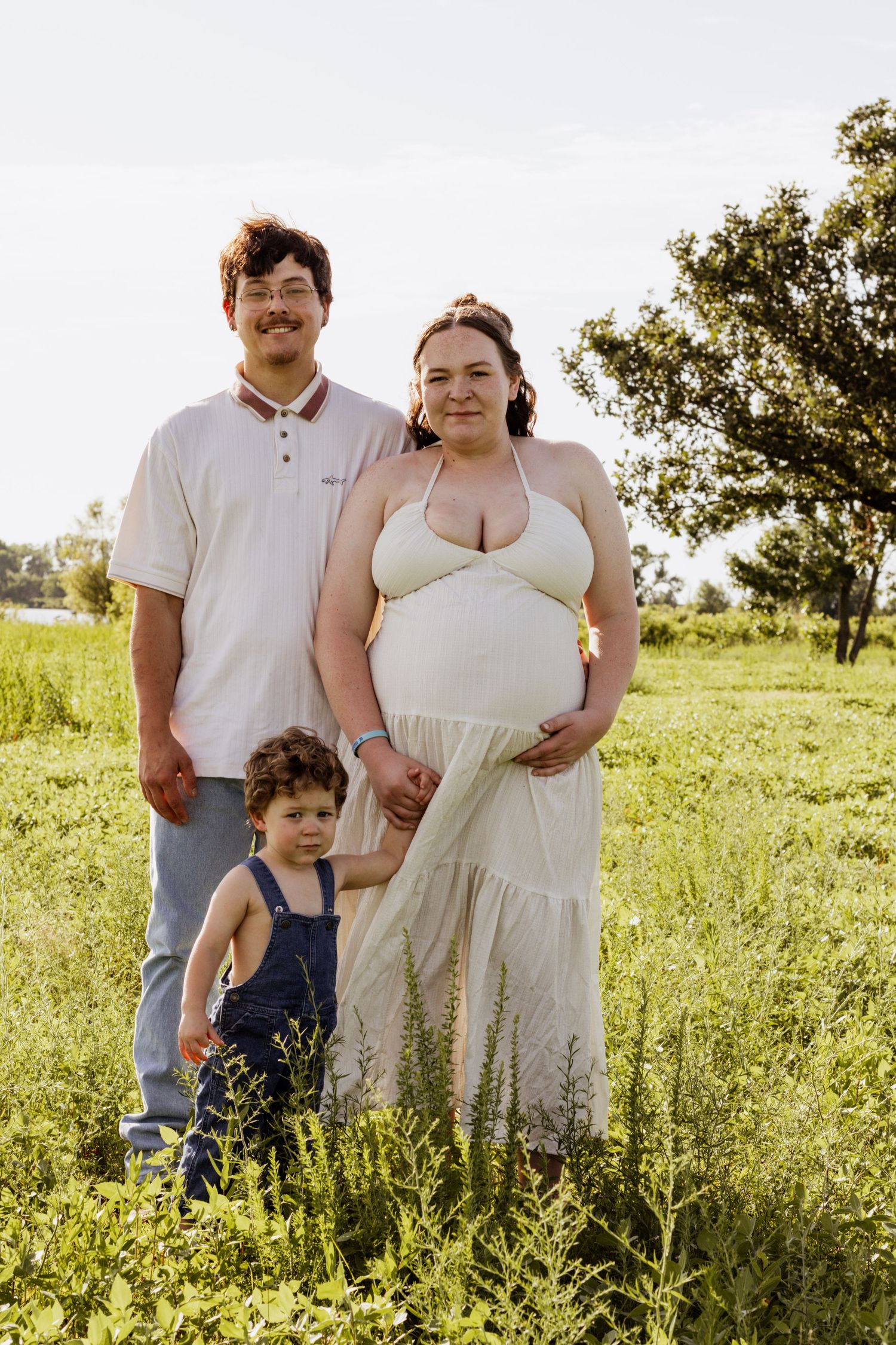 Pregnant woman in white dress poses outdoors with partner and toddler in overalls in a lush green field.