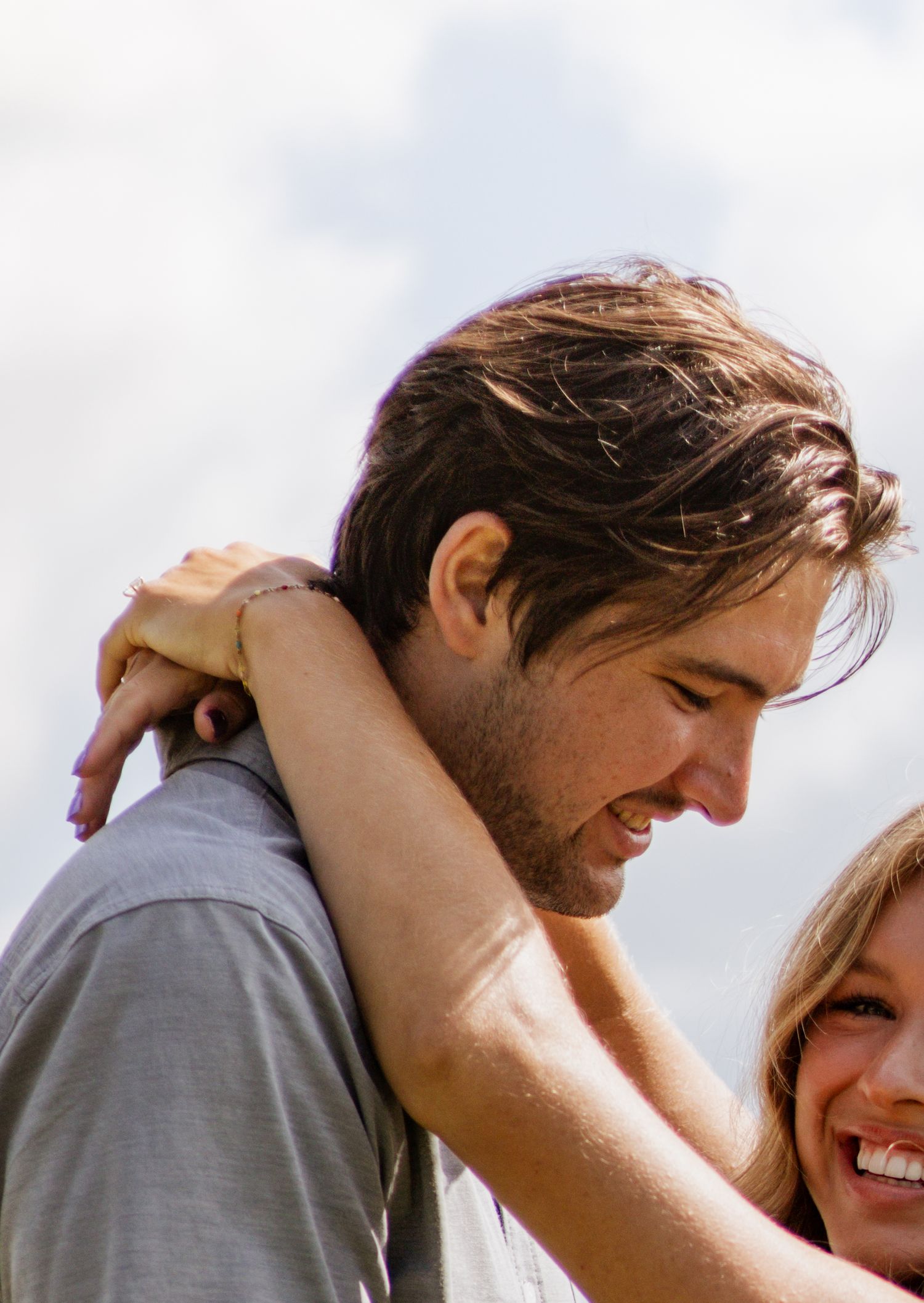 A smiling couple embraces outdoors, woman wrapping arms around mans neck, sharing a joyful moment under a cloudy sky.