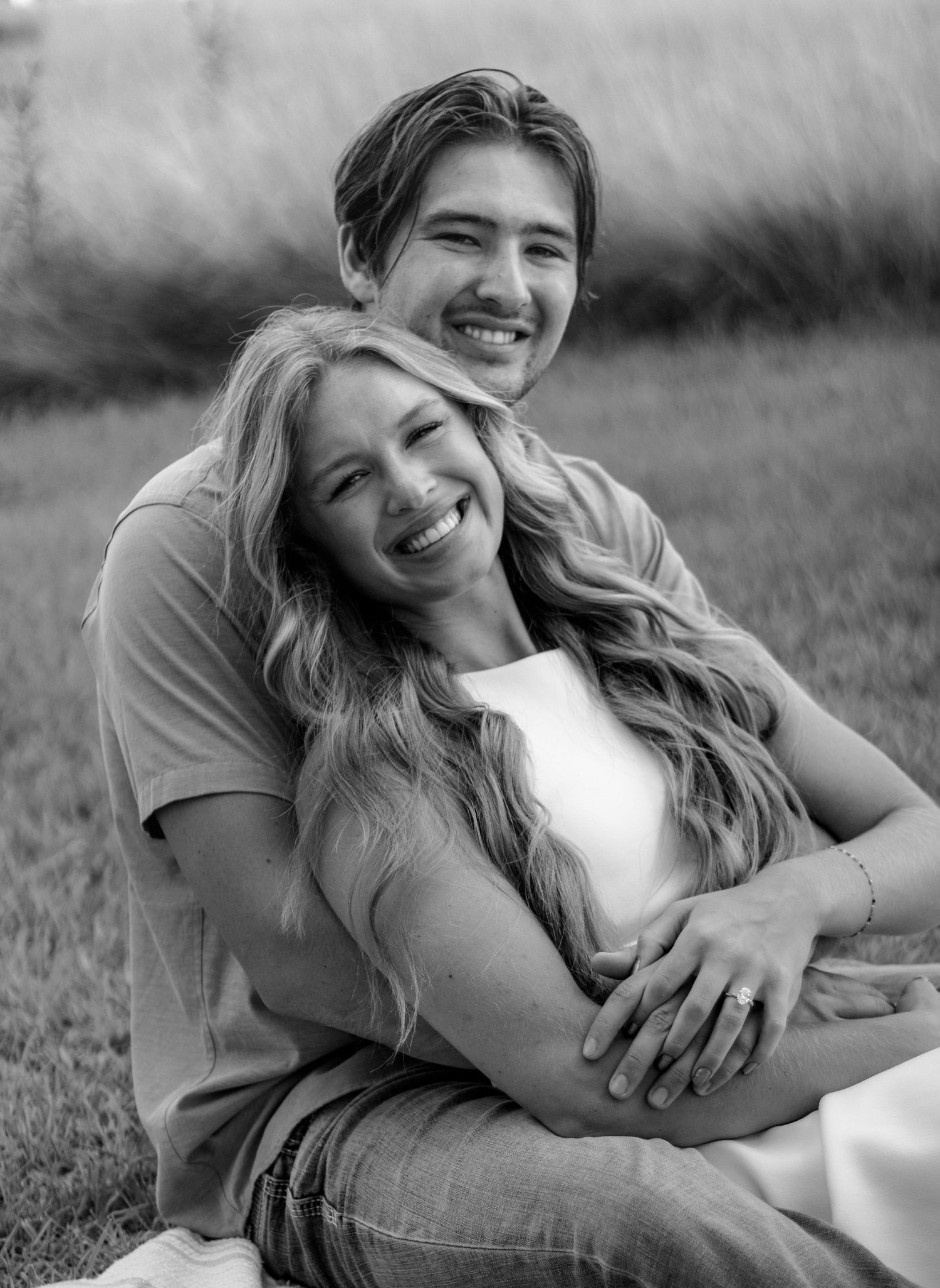 Black and white portrait of a happy couple sitting on grass, man hugging woman from behind outdoors.