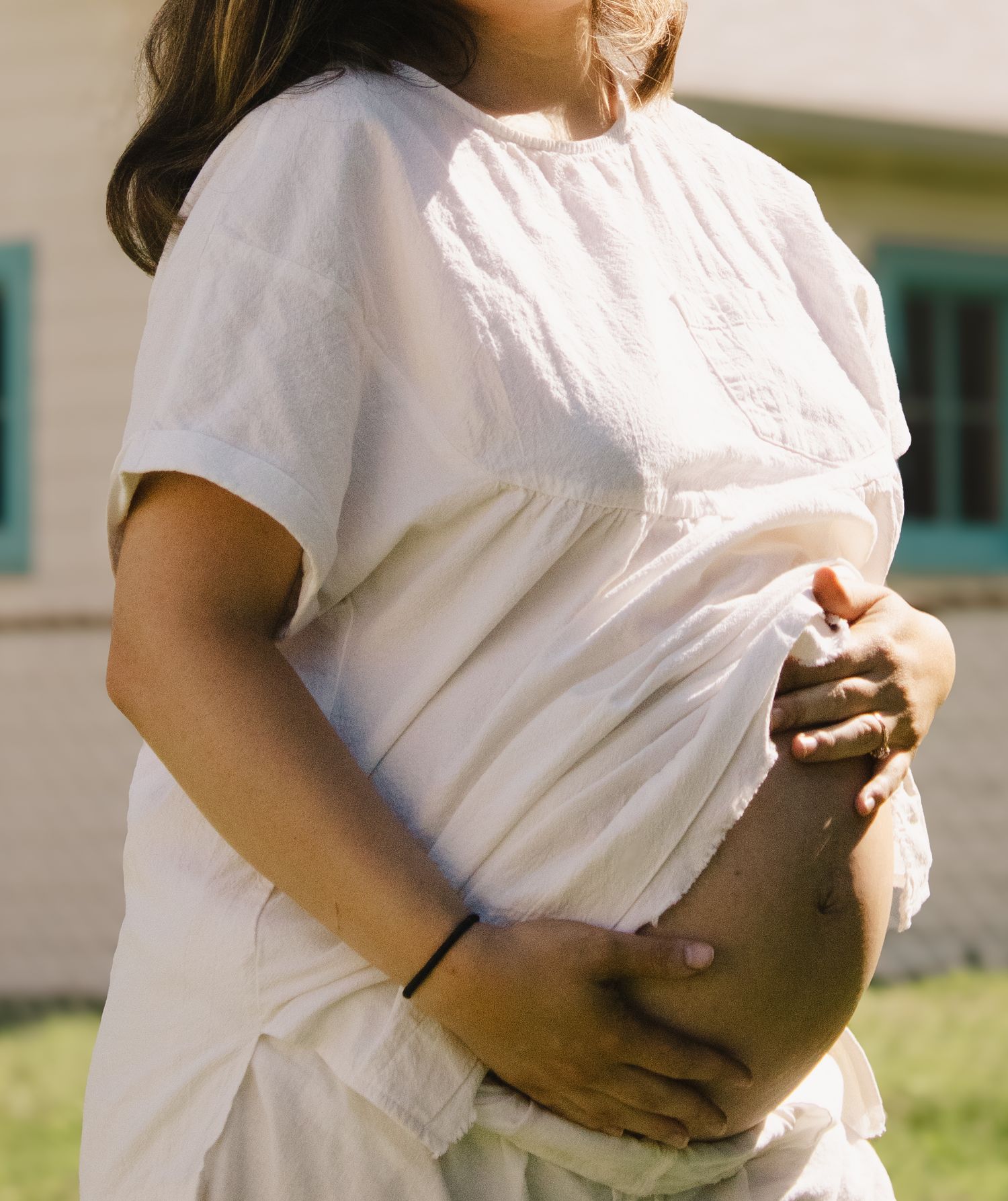 Pregnant woman in white top outdoors cradling her bare bump in sunlight.