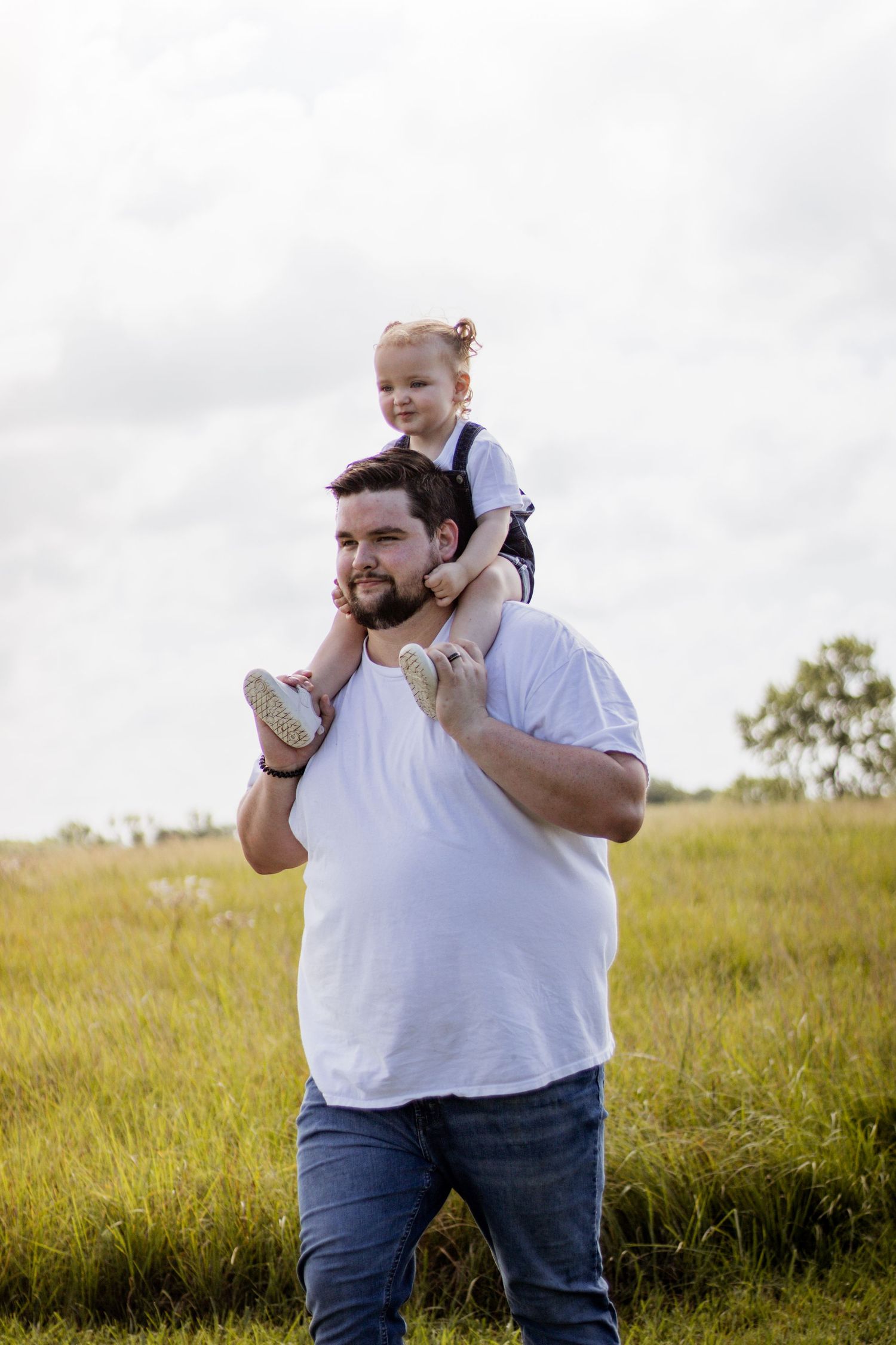 A father carries his smiling toddler on his shoulders in a grassy field, both enjoying a sunny outdoor moment.