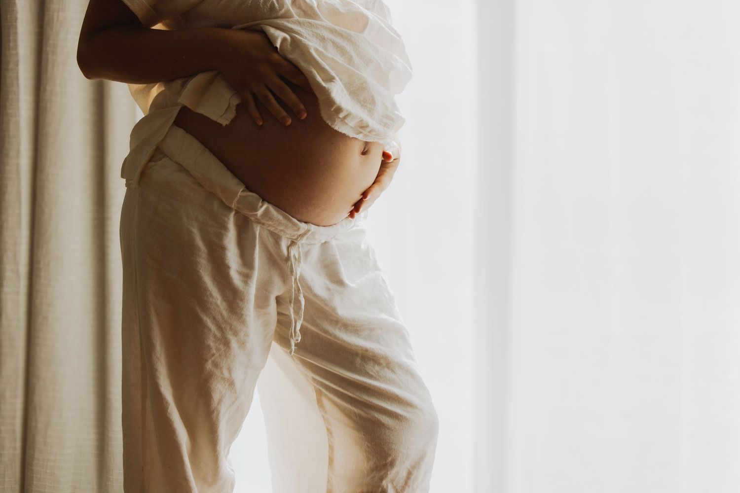 Close-up side view of pregnant woman in white outfit holding baby bump near bright window.