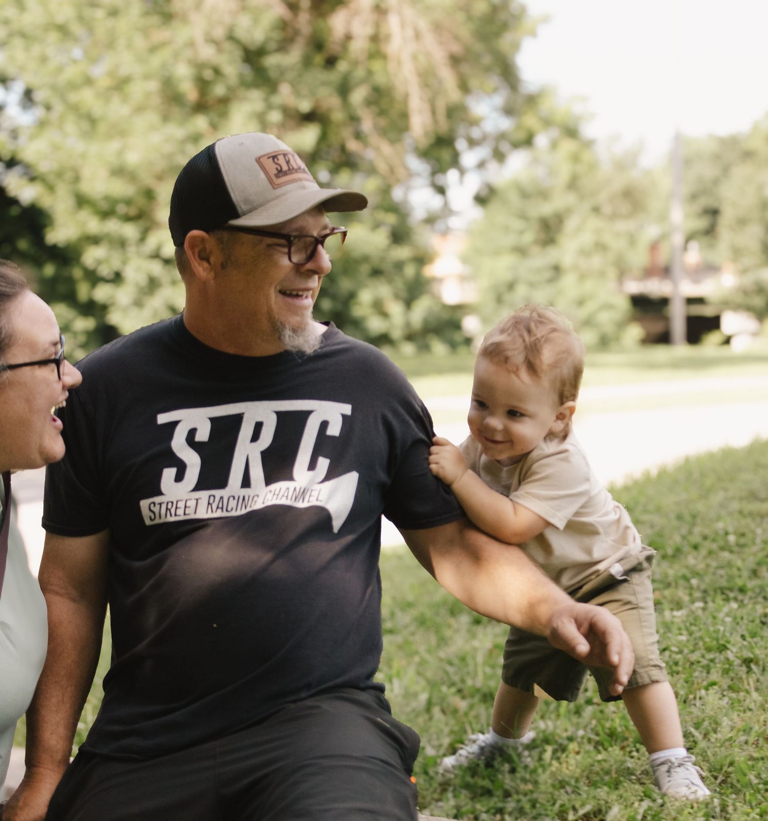 Man in SRC Street Racing Channel tee and cap laughs with woman while toddler stands on grass outdoors.