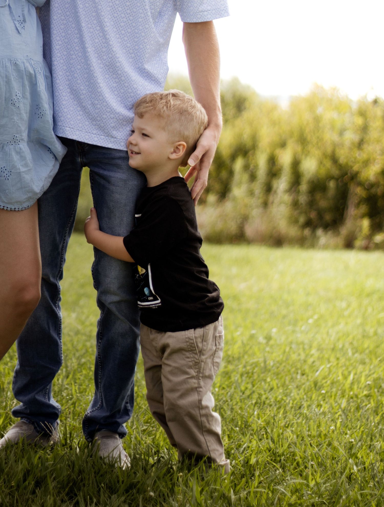 A young boy hugs an adult's legs while standing on green grass outdoors in warm sunlight.