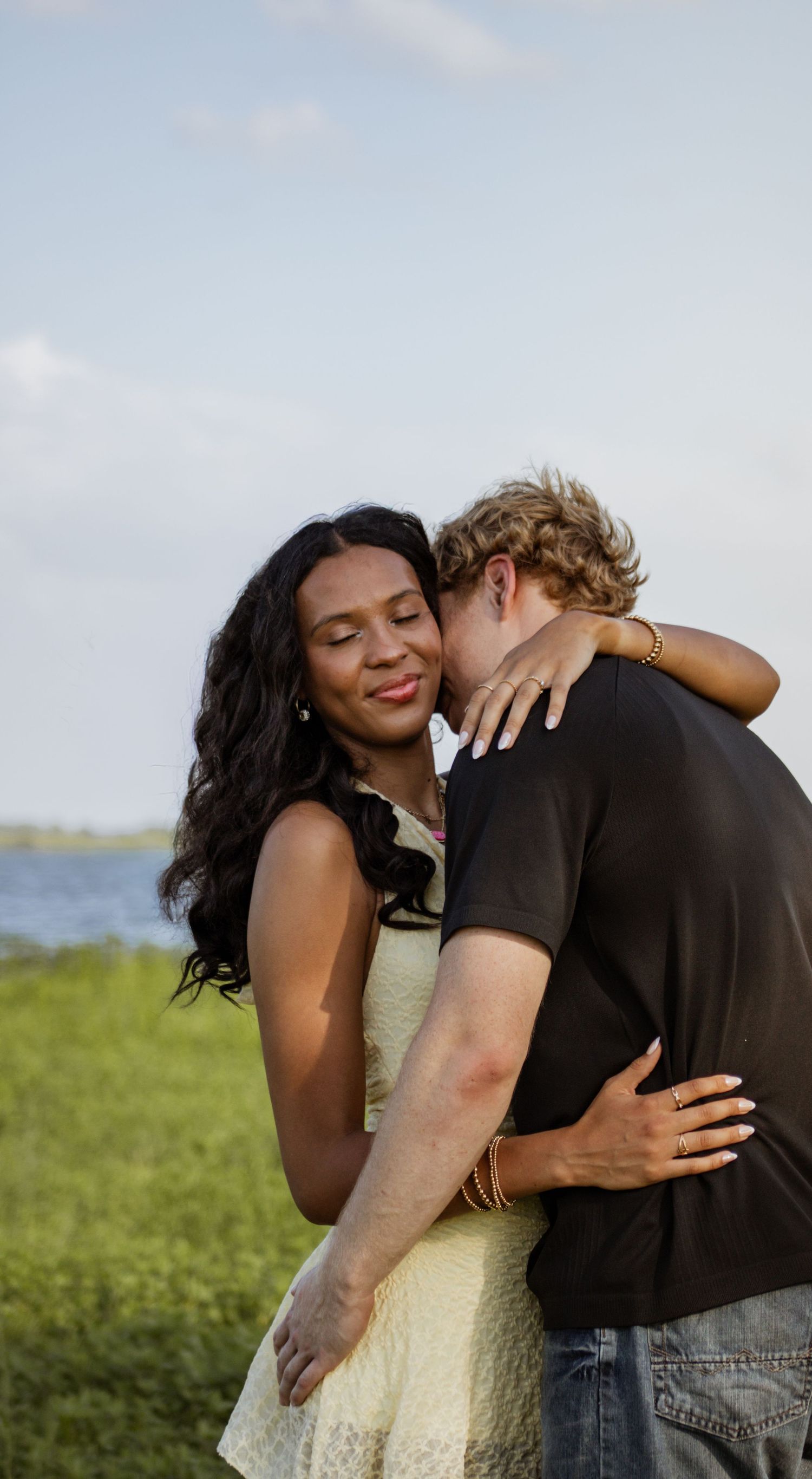 A woman with long dark hair smiles warmly while embracing a man near a scenic lakeside.